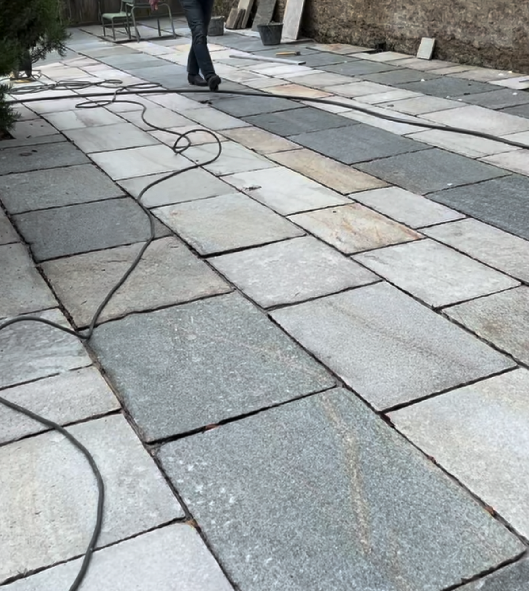 Travail de pose de pavés en pierre dans une cour, avec câbles électriques sur le sol et outils de chantier, en arrière-plan un mur en terre et des matériaux de construction.