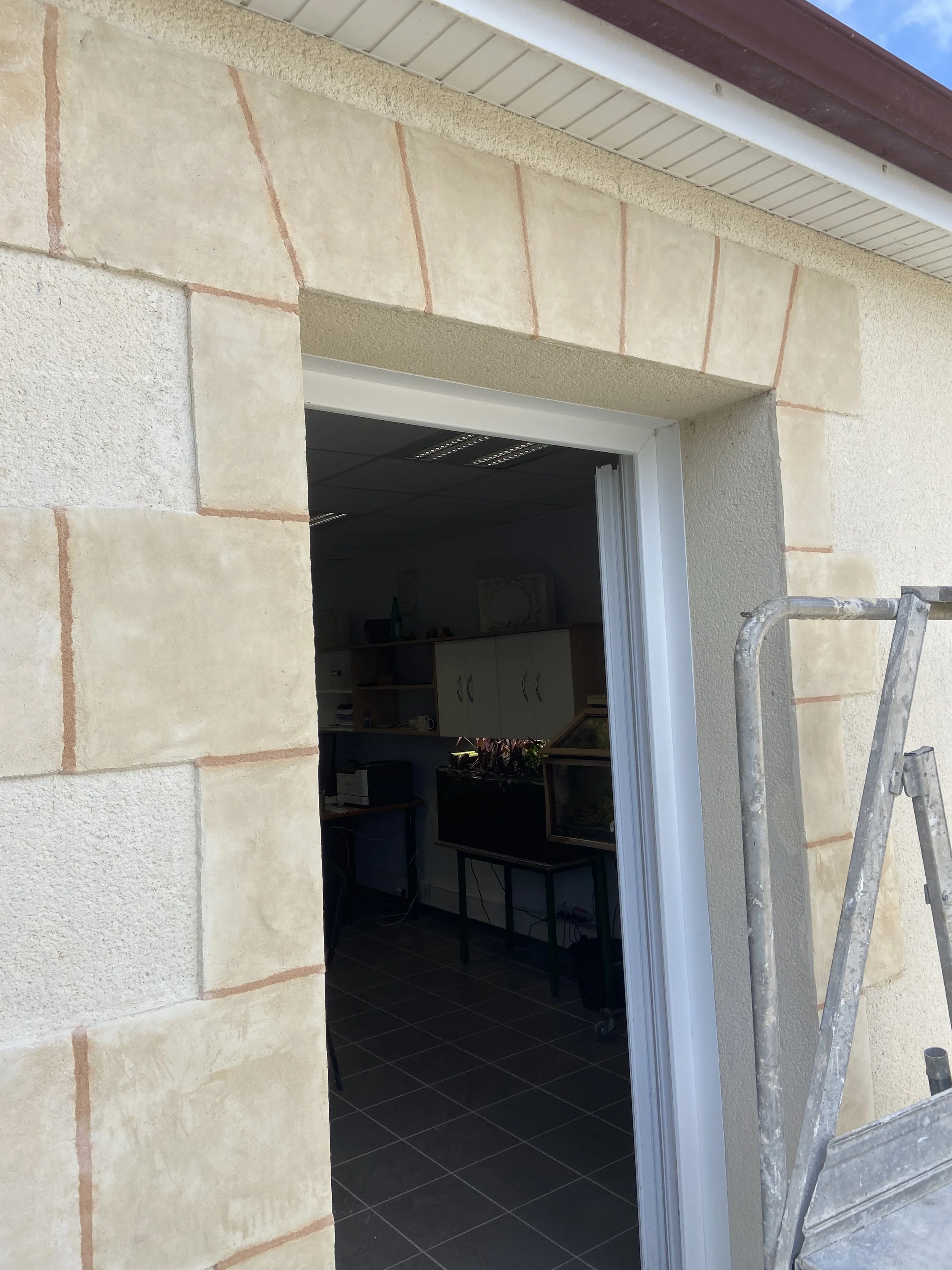 Ouverture d'une porte avec encadrement en pierre beige, mur en plâtre clair, toit en tôle, et un escalier en métal à gauche.