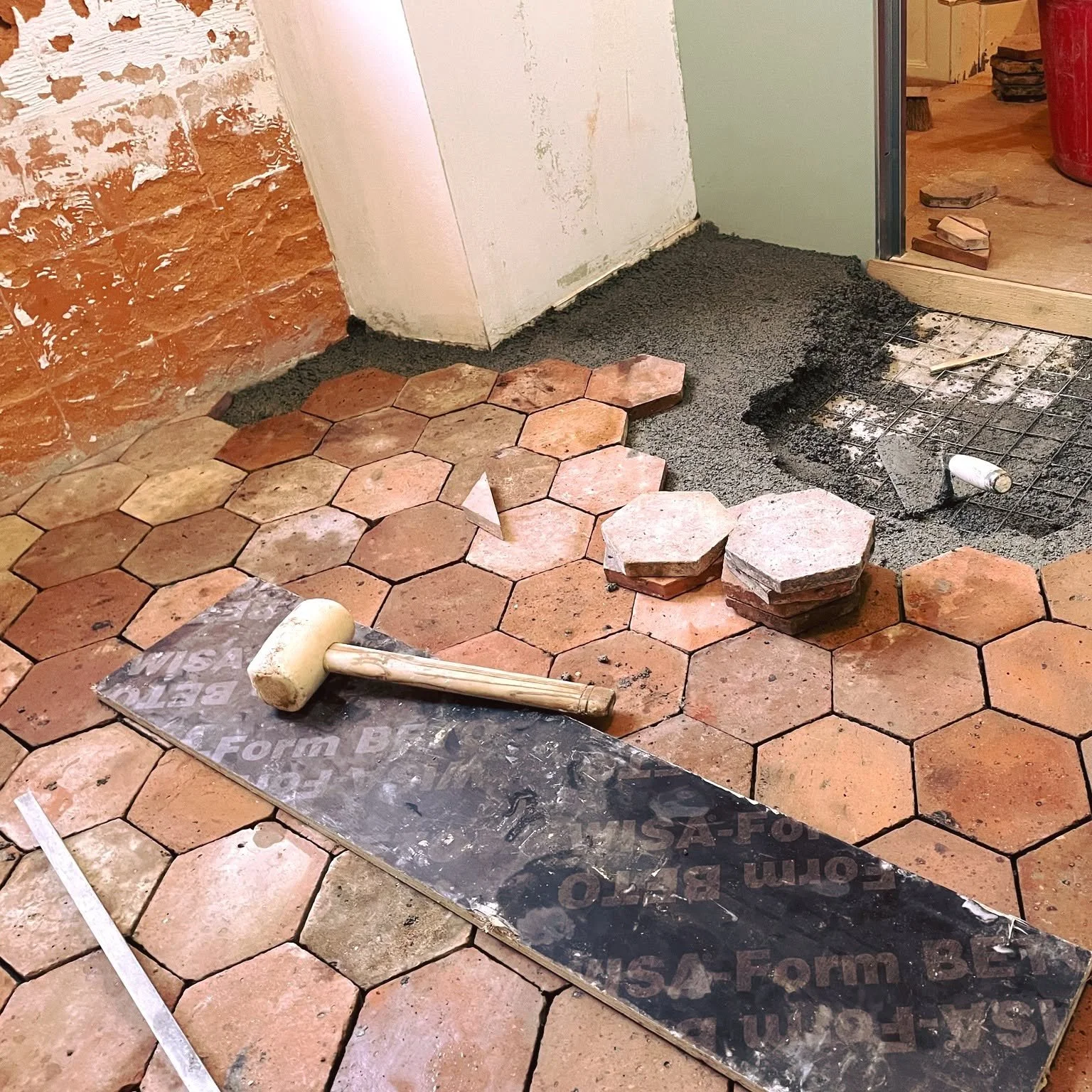 Travail de rénovation de sol avec des tuiles hexagonales en terre cuite, chape de béton en cours de pose, rouleau de joint, outils de maçonnerie, et murs en cours de finition.