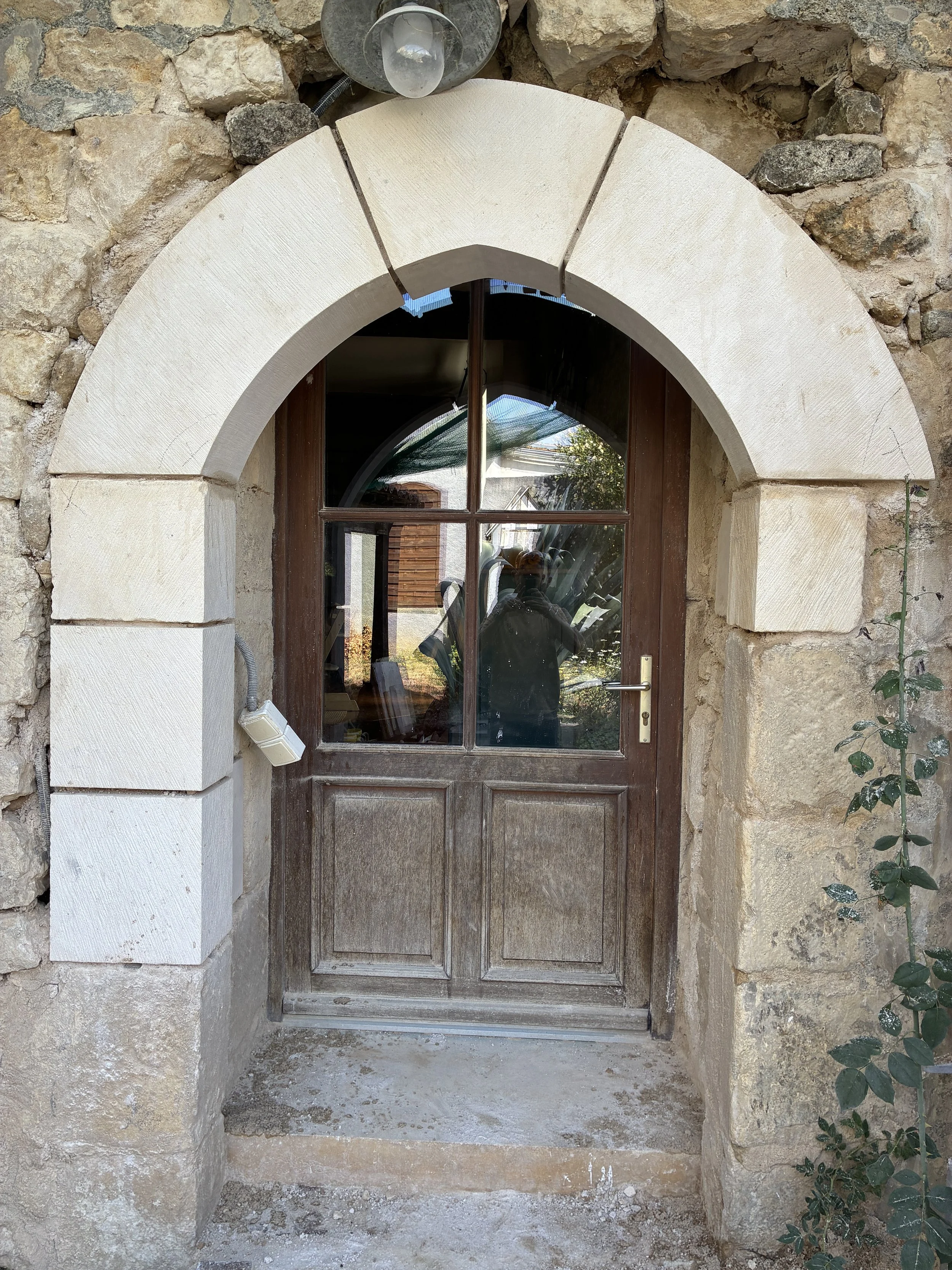 Une porte en bois avec une fenêtre en verre rectangular dans un mur en pierre. Le dessus de la porte est encadré par une arche en pierre. Il y a une lanterne fixée au mur au-dessus de la porte et un petit obstacle en pierre au sol devant la porte.