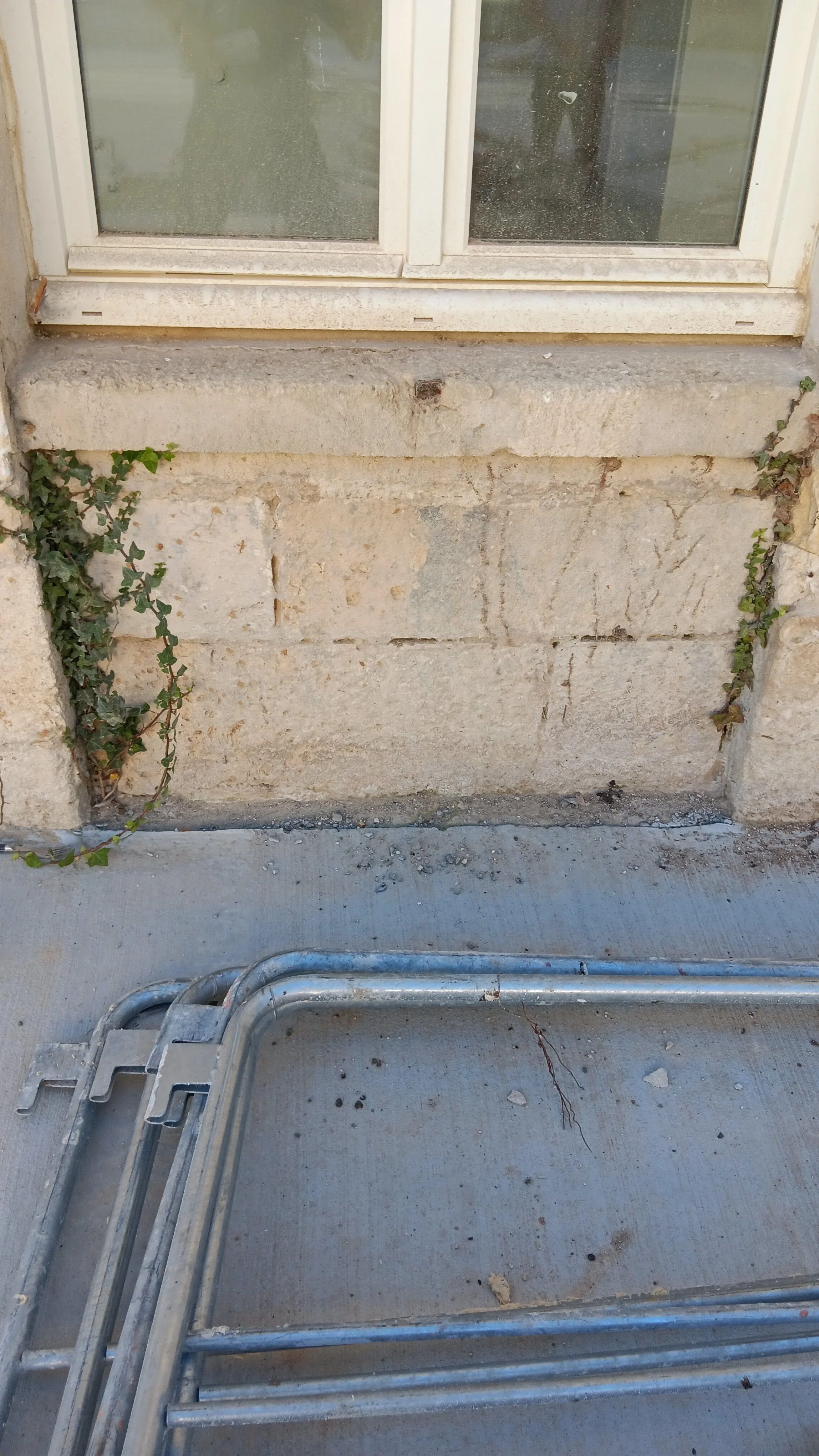 Fenêtre à double vantaux avec caisson en pierre, entourée de murs en pierre, avec des petits végétaux grimpants. Sur le sol, un cadre métallique pour fenêtres ou portes, avec un espace de chantier.