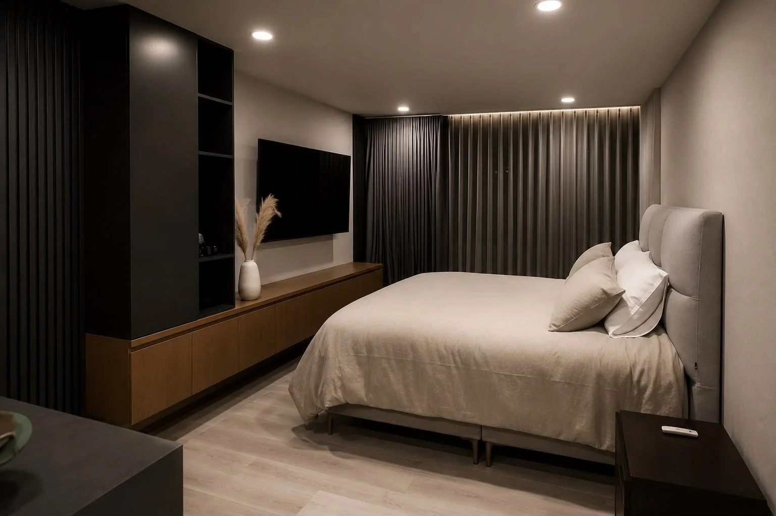 Modern bedroom with a large bed, gray upholstered headboard, white bedding, and a nightstand with a remote control. A flat-screen TV is mounted on the wall, and there is a dark curtain covering the window. A decorative vase with pampas grass is positioned on a wooden media console.