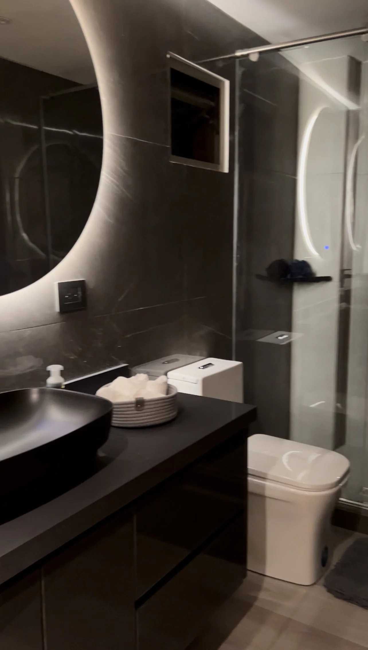 Modern bathroom with black countertop, vessel sink, and dark tiled walls. Mirror with lighting, small shelf with toiletries, visible toilet, and laundry hamper.