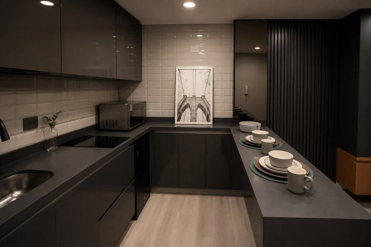 Modern kitchen with black cabinets, beige subway tile backsplash, and a gray countertop. Decor includes a framed black and white bridge drawing, a small vase with a green plant, and white tableware set on the counter. Recessed lighting illuminates the space.