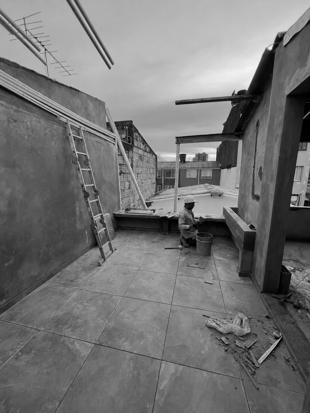 Construction worker kneeling on a tiled rooftop terrace, working on a low ledge, with construction tools and materials scattered around. A ladder leans against the adjacent wall, and building structures are visible in the background under a cloudy sky.
