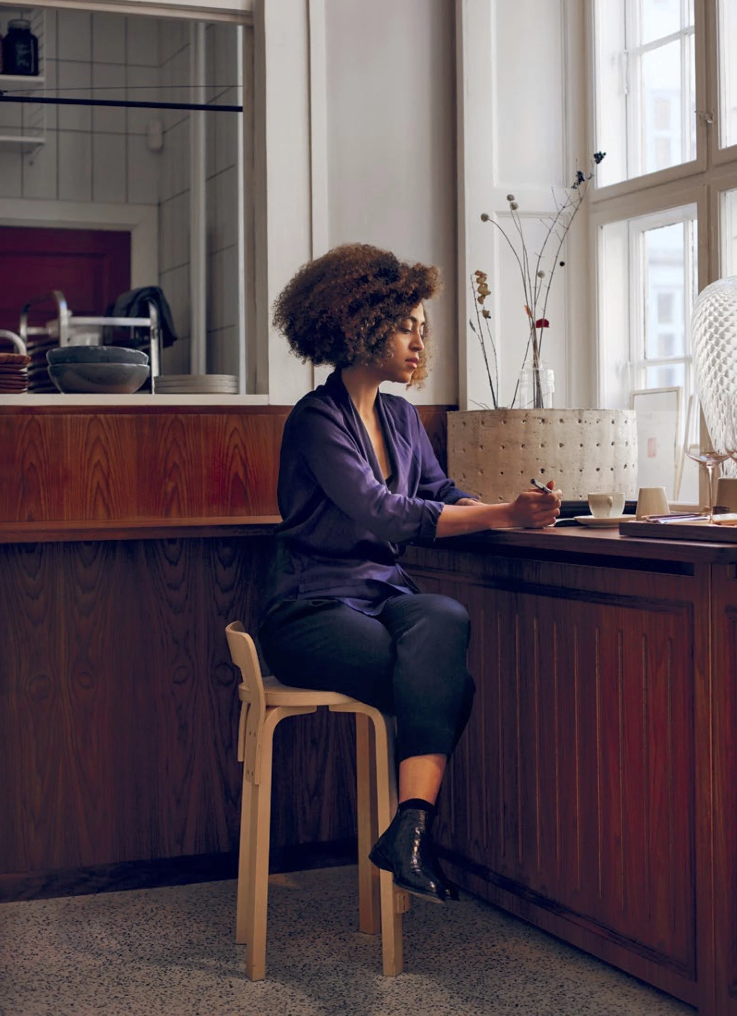 Angela sitting at a wooden bar counter with a window behind her. She appears to be holding a spoon or utensil, with dishes, cups, and a teapot on the counter. The setting looks like a cozy cafe or restaurant interior.