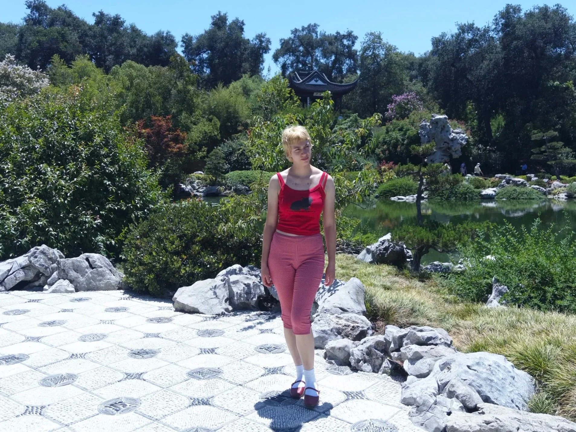 A woman standing on a stone pathway in a lush garden with trees, shrubs, rocks, and a pond; a traditional East Asian style pavilion is in the background, under a clear blue sky.