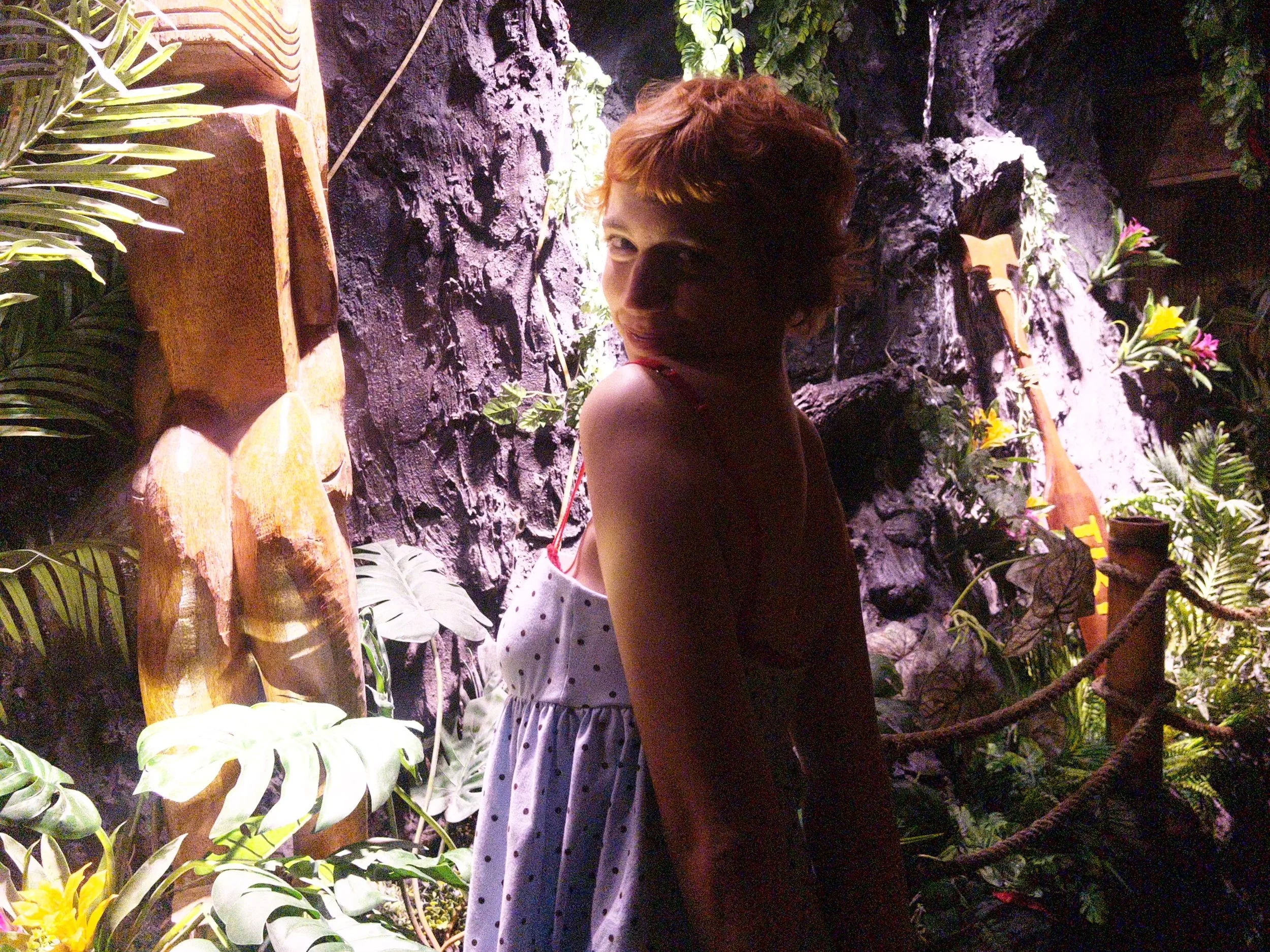 A woman with short reddish hair in a polka dot dress looking over her shoulder in a lush, tropical-themed environment with rocks, plants, and flowers.
