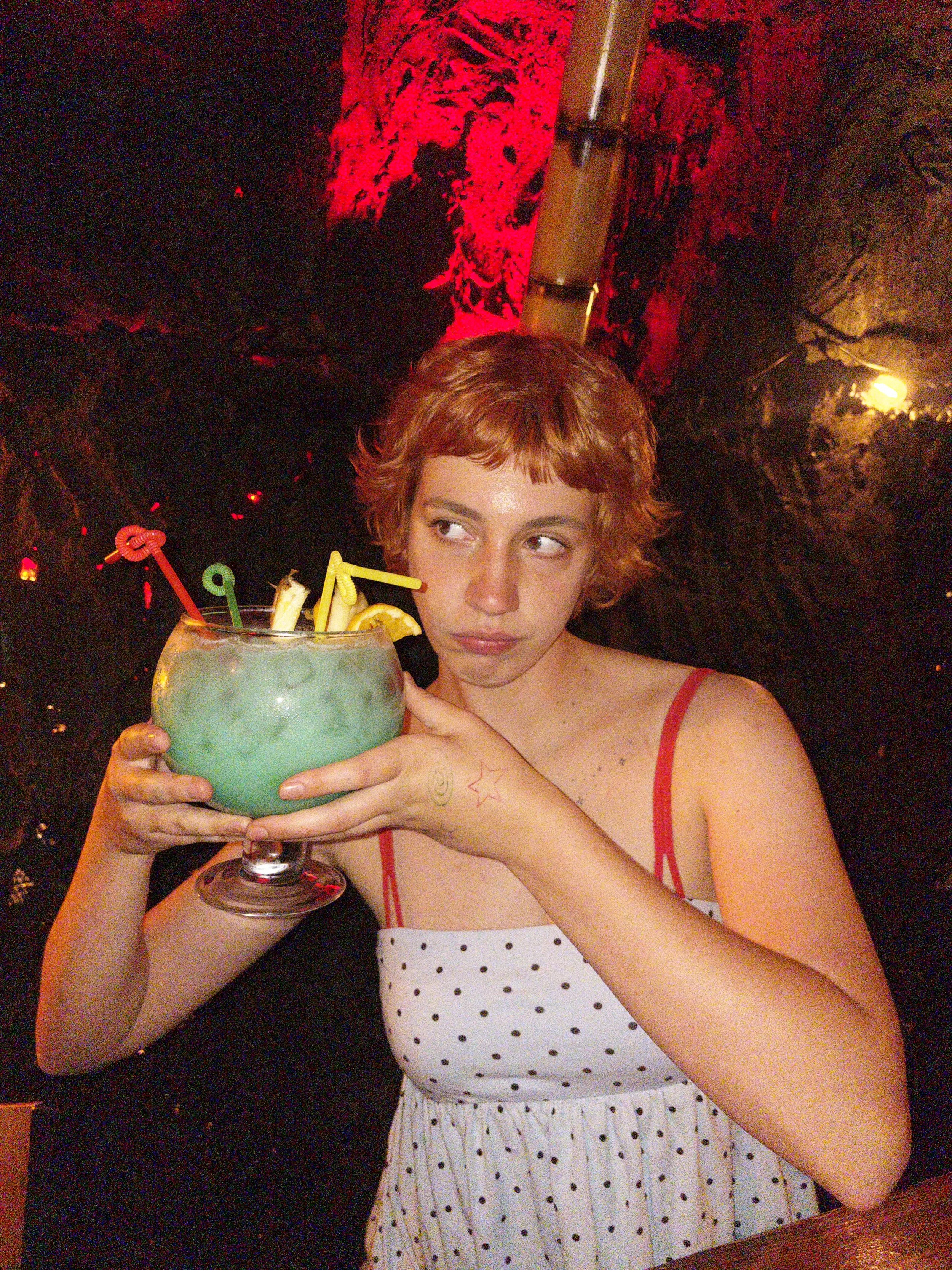 A young woman with short, curly red hair holding a large blue cocktail with lemon slices and colorful stirrers in a dimly lit setting with red and yellow lighting.