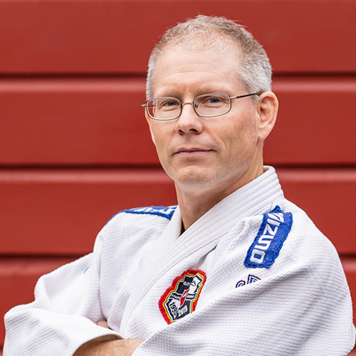 A man wearing glasses and a white martial arts gi with a patch, standing in front of a red wooden wall.
