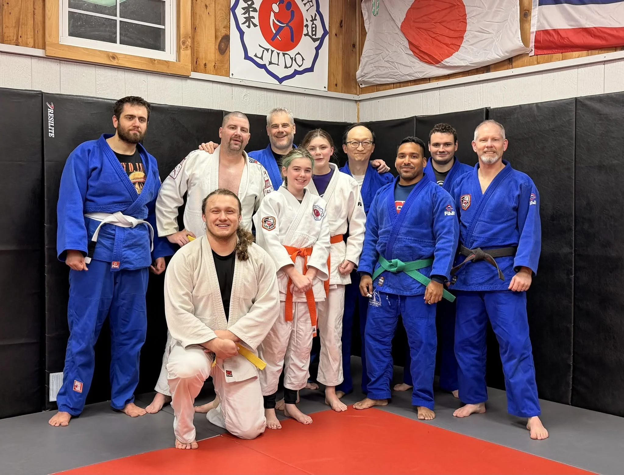 Group of judo practitioners in martial arts uniforms standing together on a mat in a dojo, smiling, with Japanese flags and dojo banner hanging on the wall behind them.