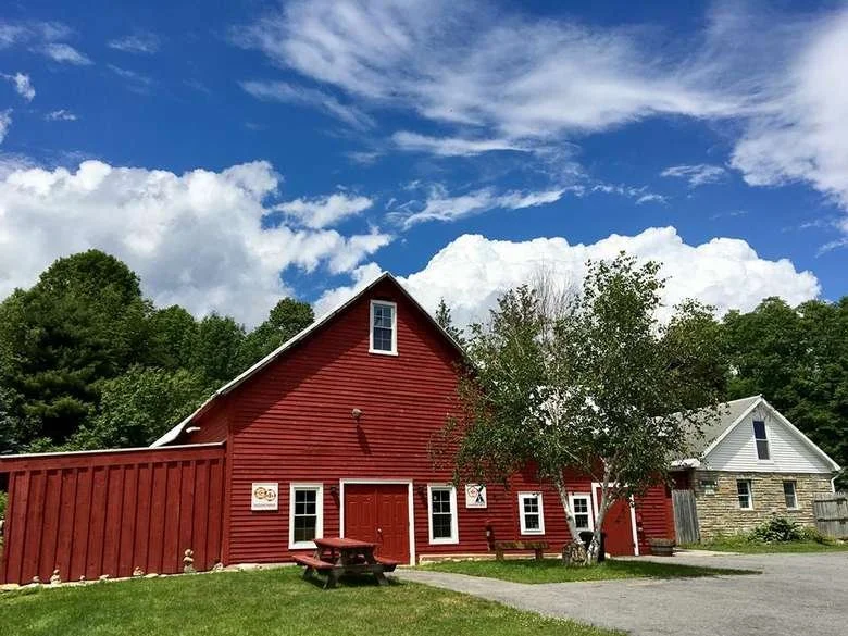A red barn with white window frames and a small door, situated next to a grey and white building, with a gravel driveway, green grass, and a large tree in the foreground under a partly cloudy sky.