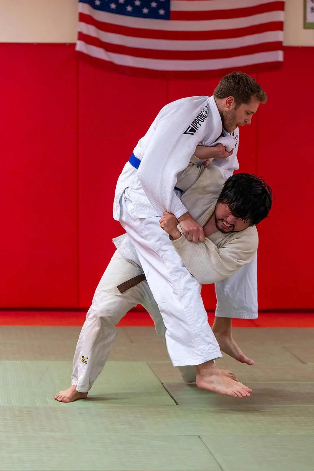 Two men practicing Brazilian Jiu-Jitsu; one is in a white gi and the other in a beige gi, engaged in a grappling move in a dojo with a red padded wall and an American flag in the background.