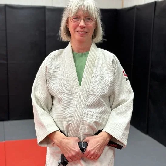 A woman with gray hair, glasses, and a green shirt underneath a white martial arts gi, standing on a red and black mat in a martial arts dojo.
