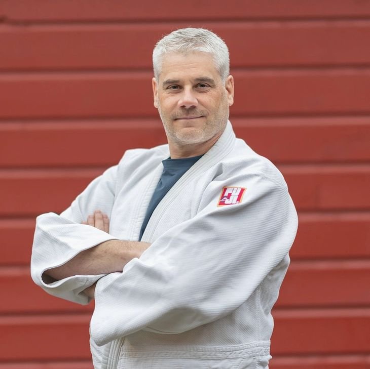 A man with gray hair dressed in a white martial arts gi standing in front of a red wooden wall, arms crossed, smiling confidently.