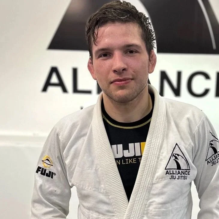 Young man with dark hair and a slight smile, wearing a white martial arts gi with patches, standing in front of a wall with the word 'ALLIANCE' in large letters.