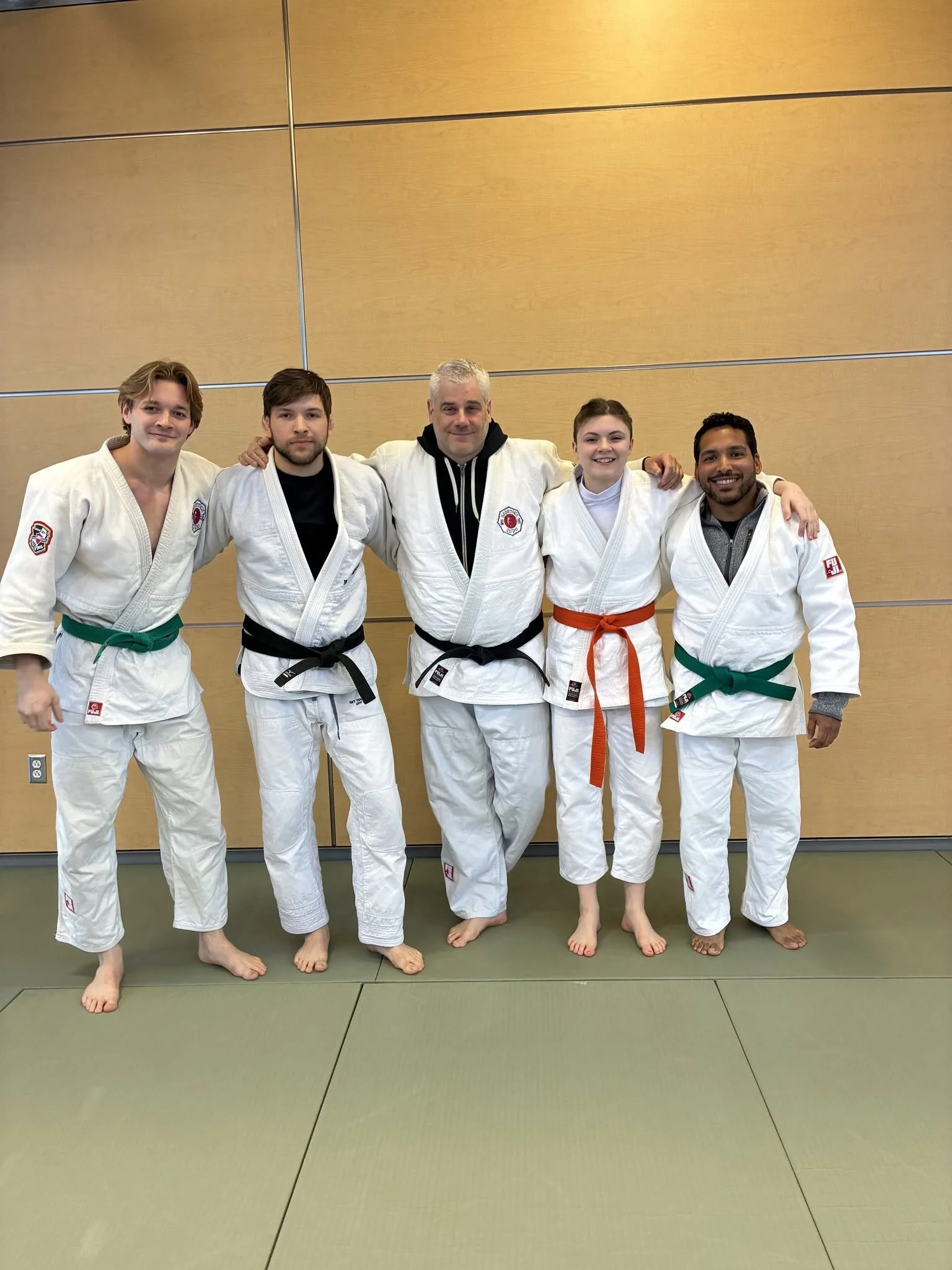 Five people in martial arts uniforms standing together in a dojo with their arms around each other, smiling.