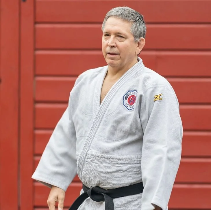 A man in a white martial arts gi with a black belt, standing outdoors against a red wooden wall.