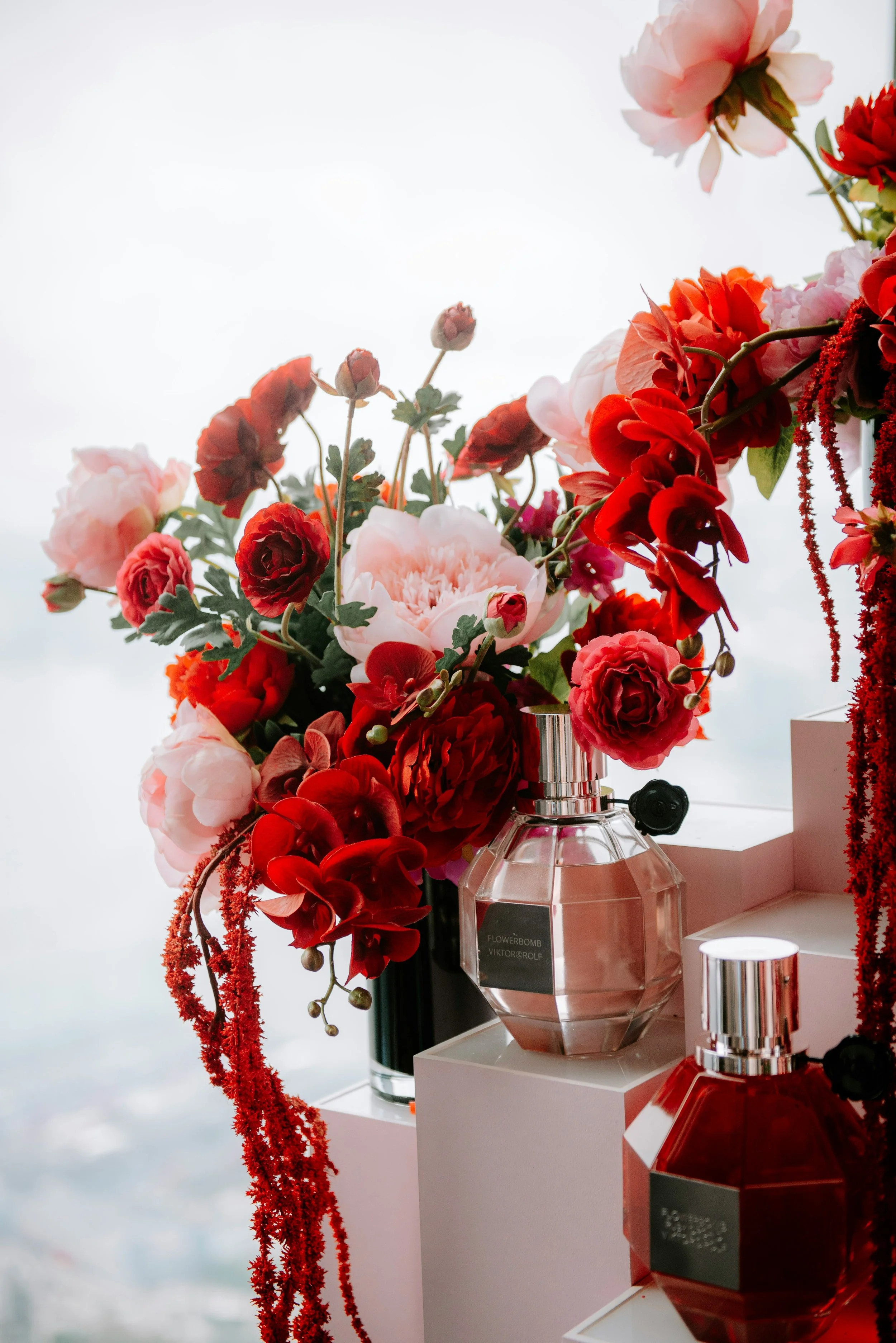 A close-up of a floral arrangement with pink, red, and white flowers in vases accompanied by bottles of perfume, set against a blurred background.