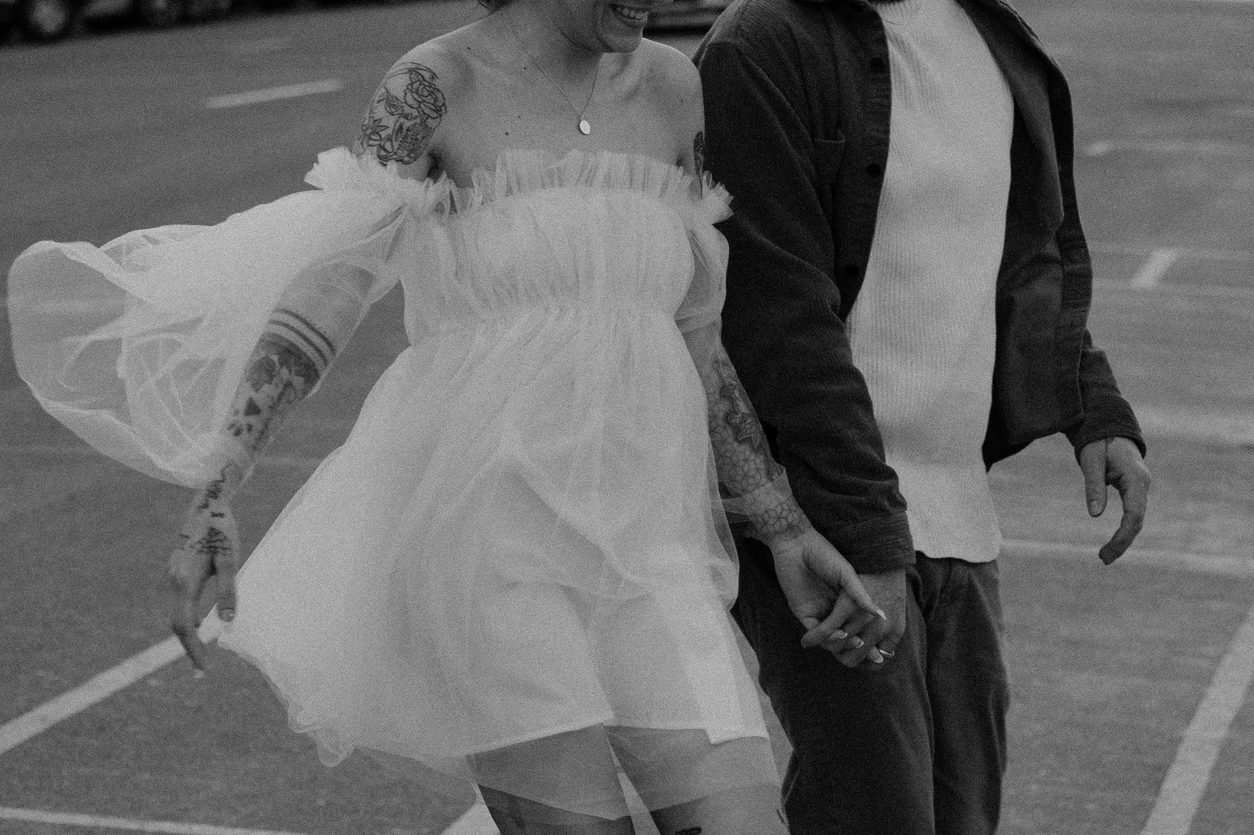 A candid black-and-white photo of a couple skating hand-in-hand at an outdoor rink. The woman is dressed in a white, off-the-shoulder dress with lace detail and has tattoos on her arm. The man is wearing a white shirt and a dark jacket.