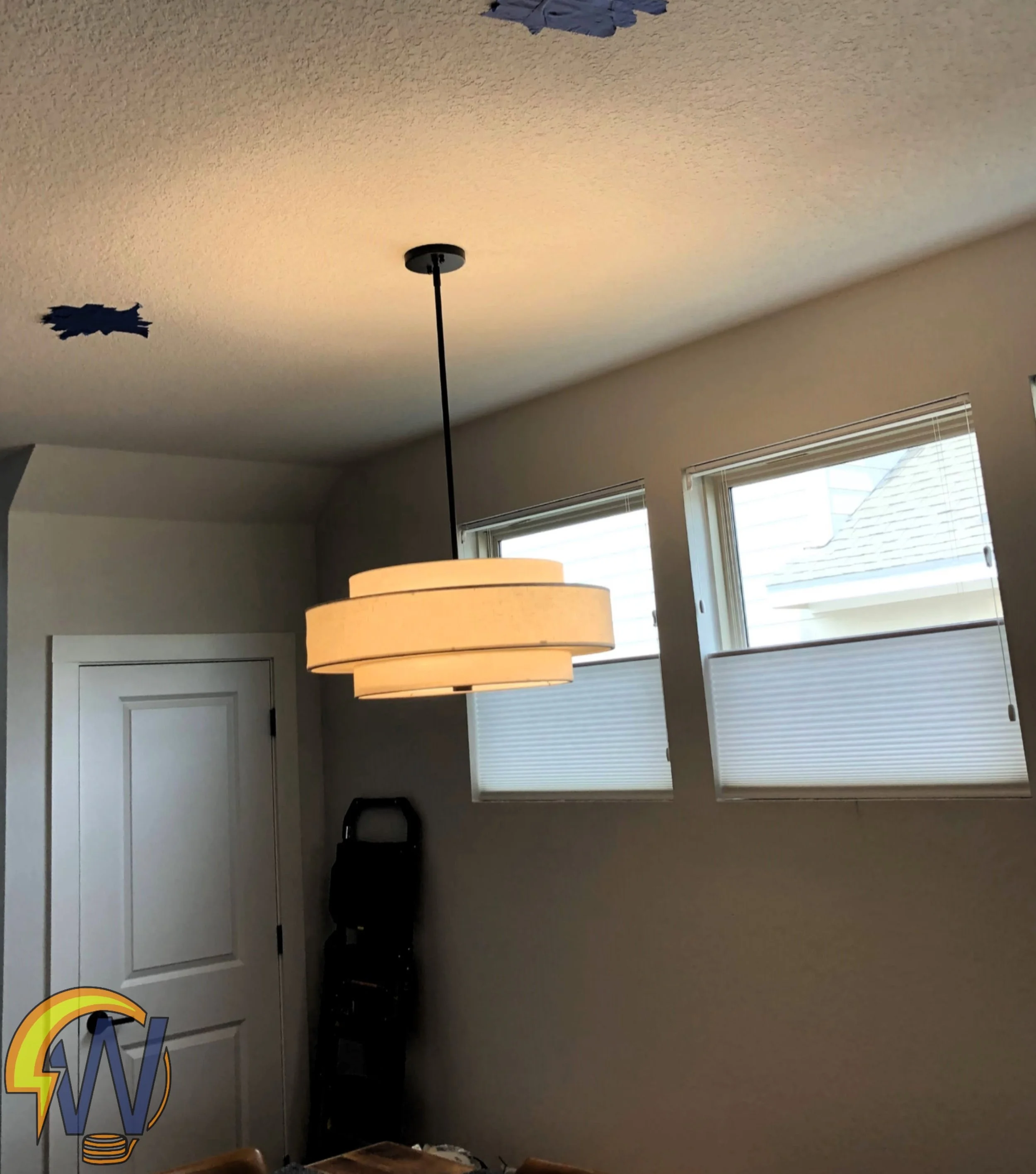 A room with beige walls and a ceiling, featuring a hanging black pendant lamp and two windows with white blinds, one window shows a roof with shingles outside.