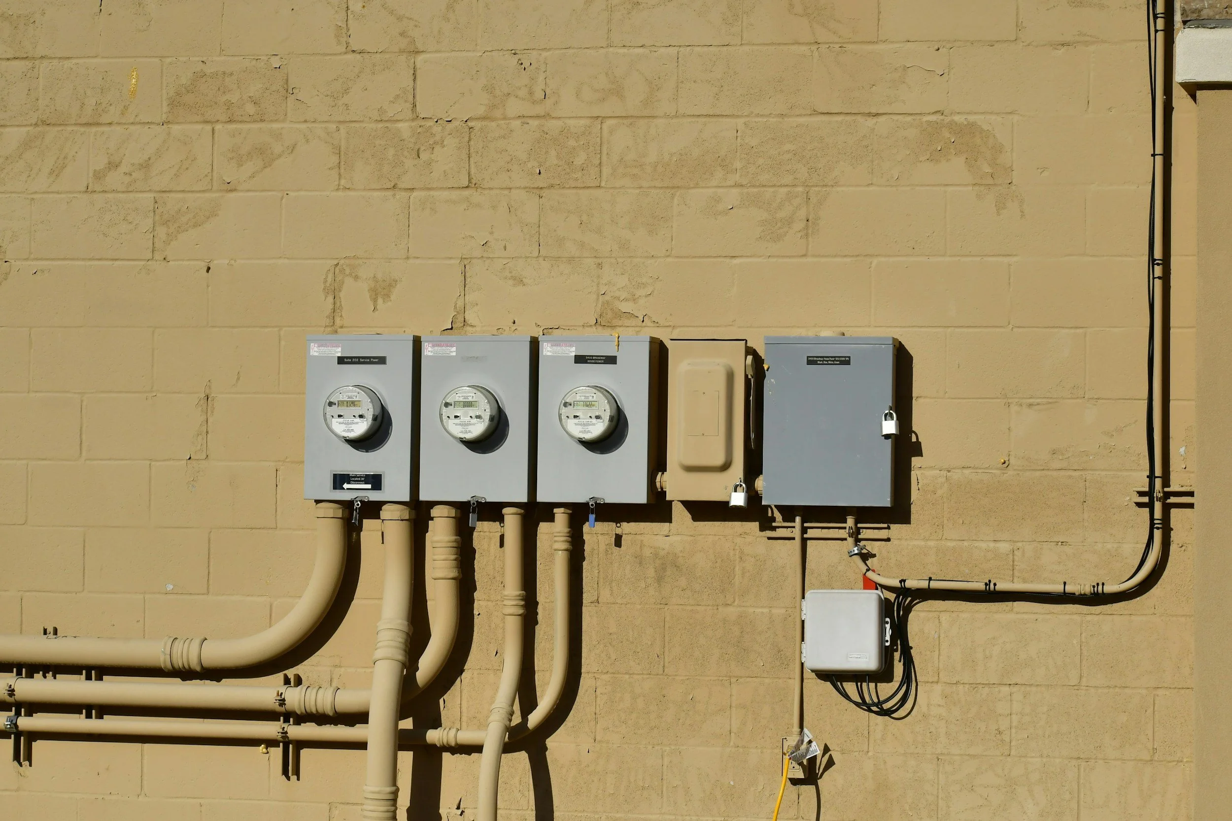 Four electric meters mounted on a beige brick wall, with pipes and a locked electrical box below and to the right of the meters.