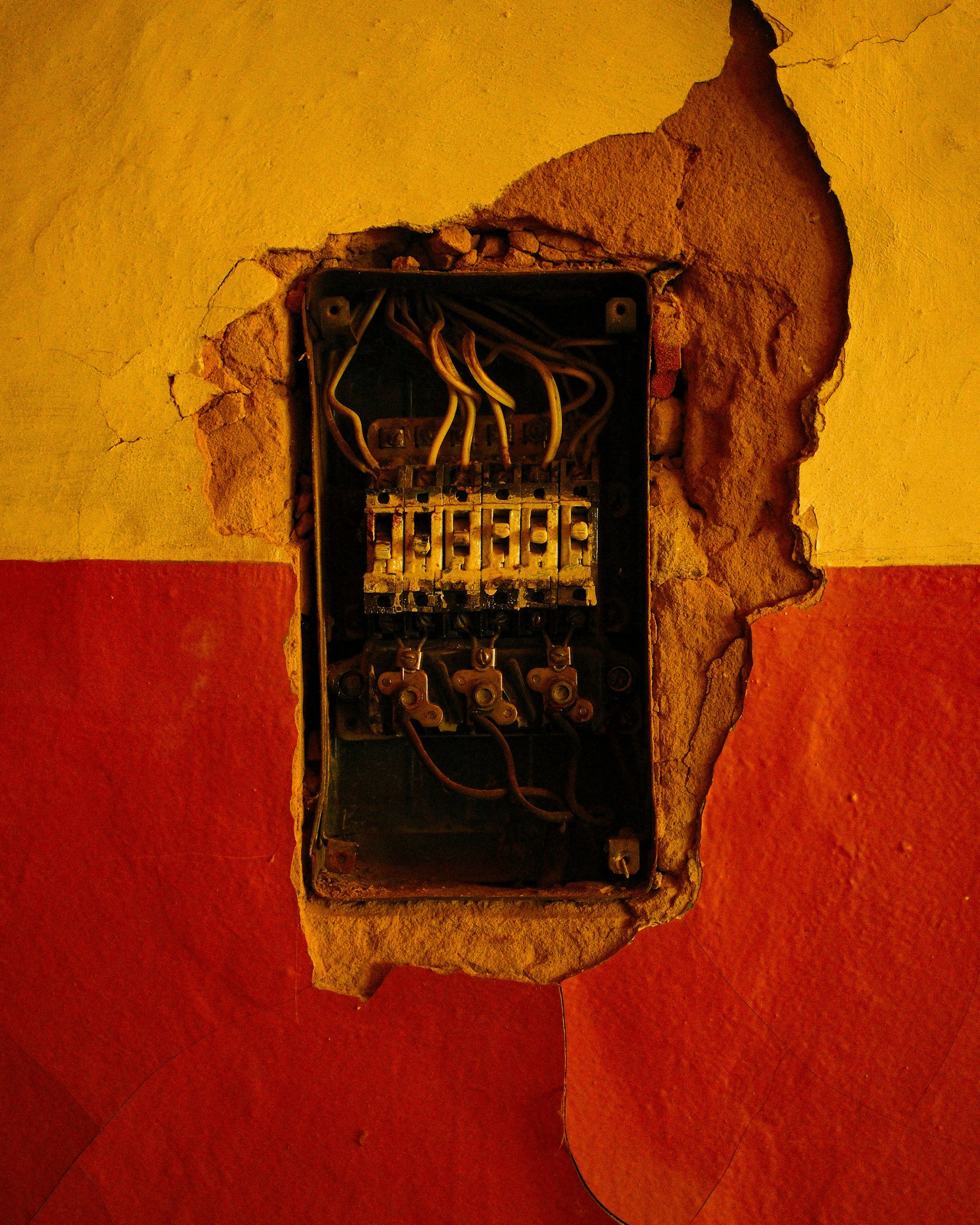 An open junction box with exposed electrical wires embedded in a wall with yellow and red paint, and a cracked section of the wall surrounding it.