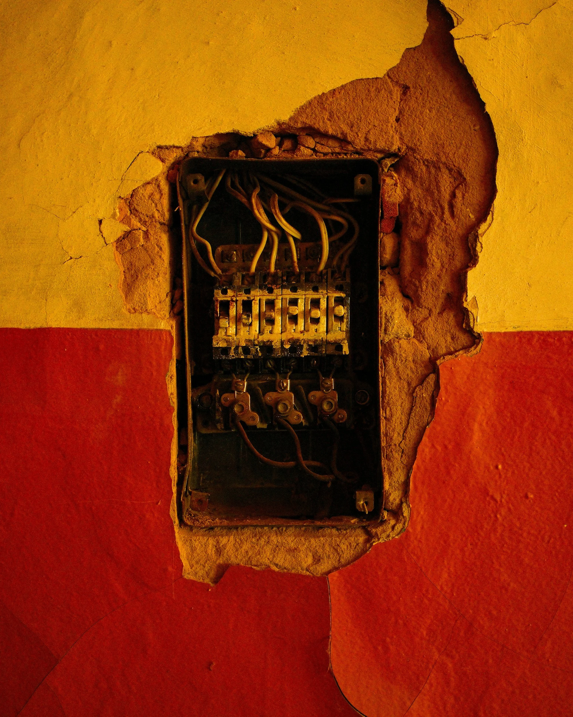 An open electrical panel with exposed wiring mounted on a wall with peeling paint, revealing brick underneath.