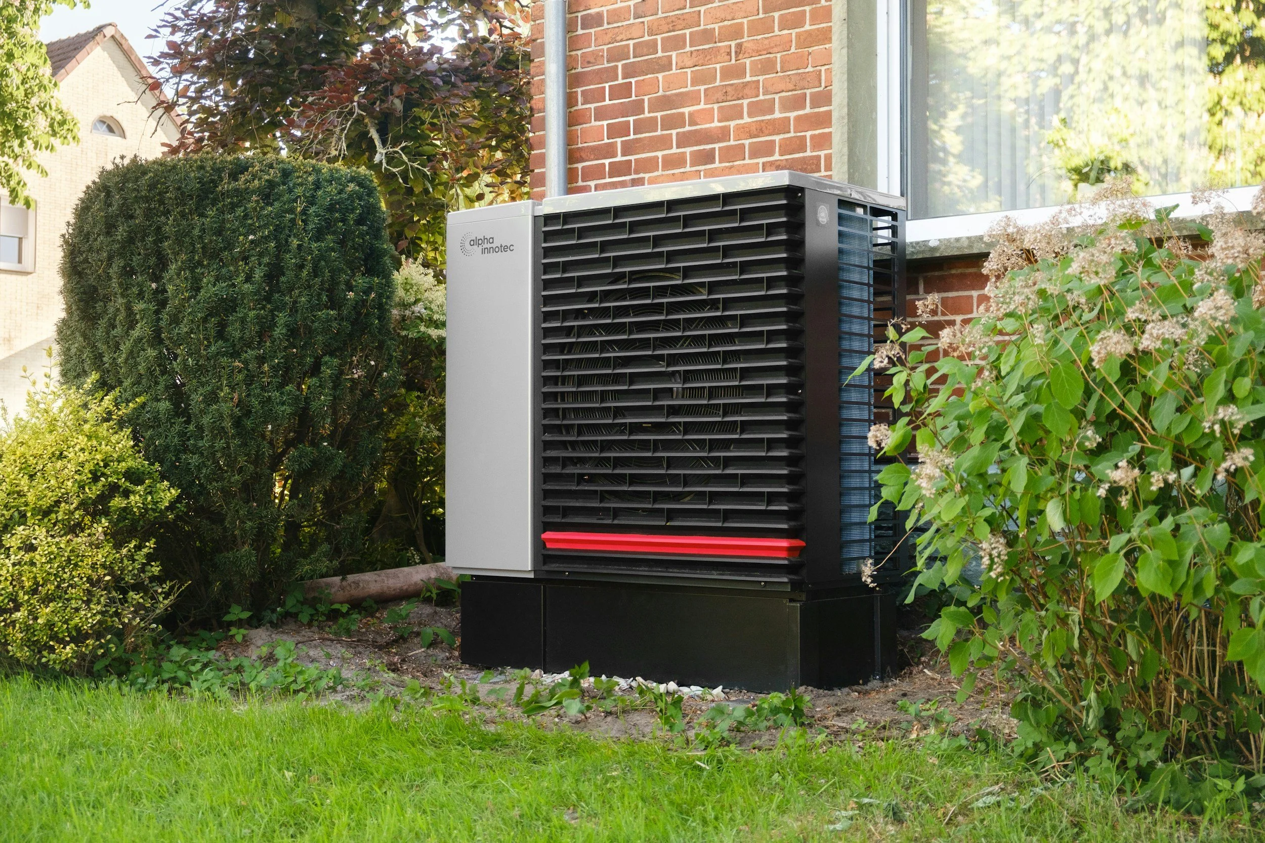 Outdoor air conditioning unit installed near a brick house wall surrounded by bushes and greenery.