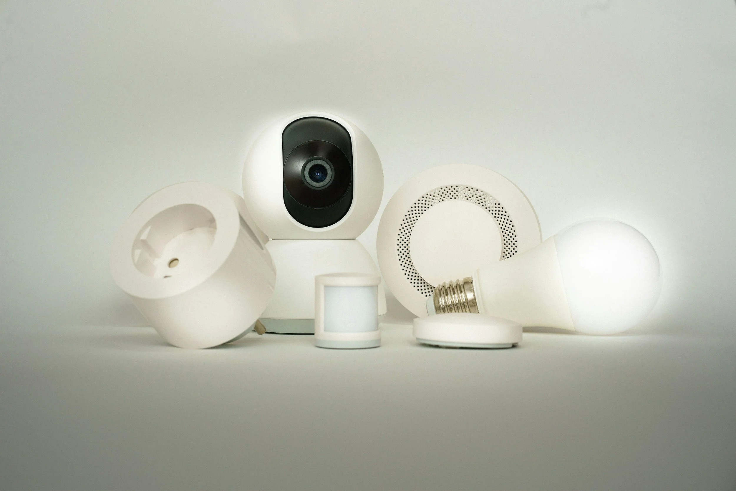 Various white smart home devices and lightbulbs arranged on a table, including a security camera, speakers, and lightbulb.