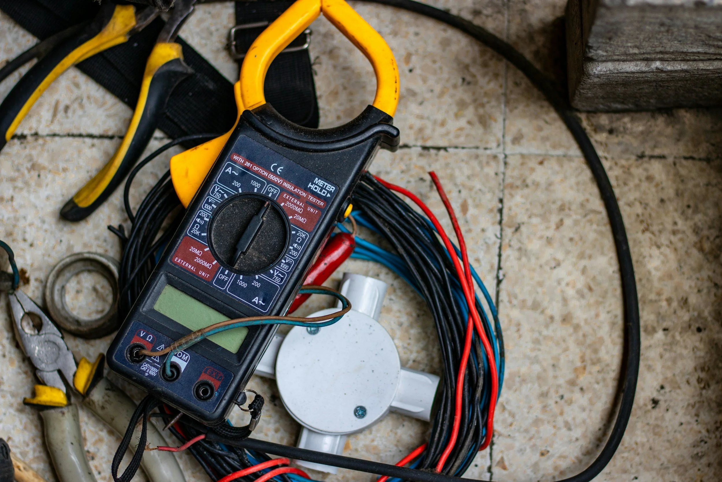 A digital multimeter resting on a tiled floor with various tangled wires, a white circular socket, and stray tools.