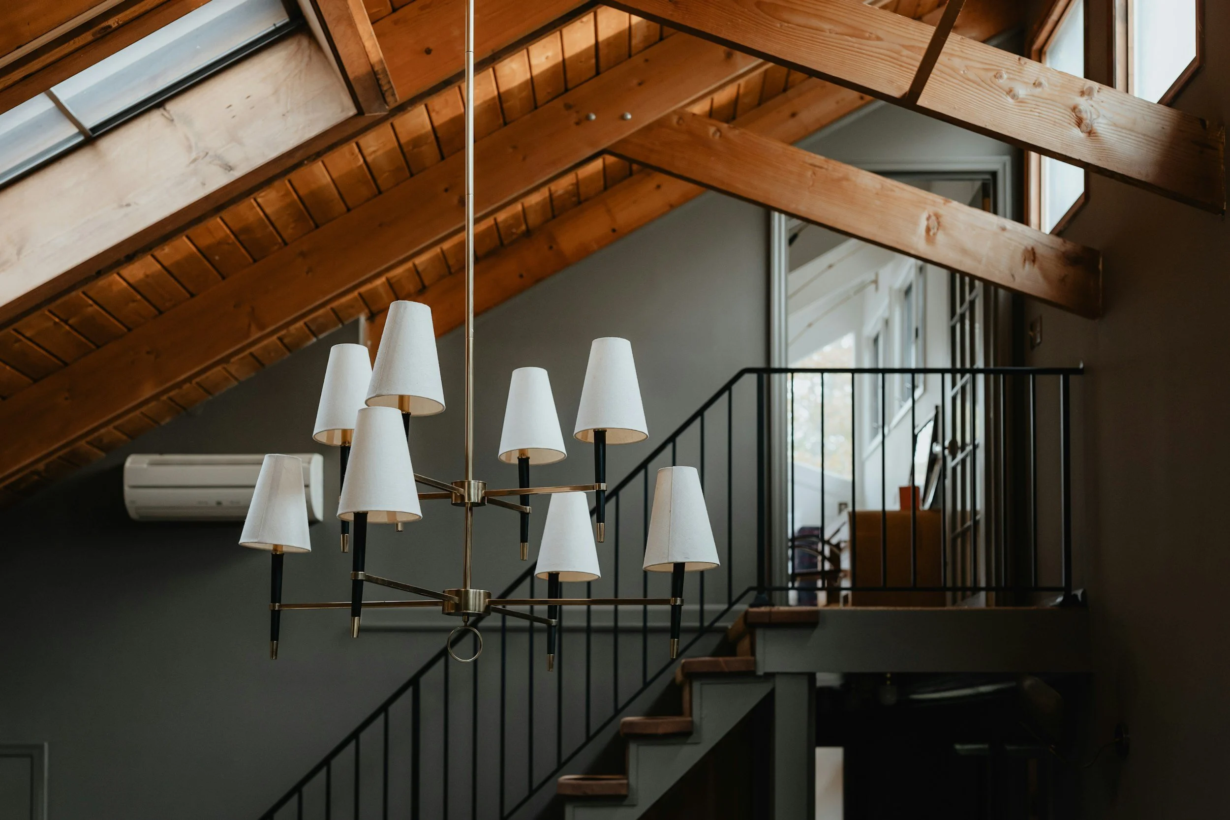 A room with wooden vaulted ceilings, a chandelier with multiple white lampshades, a staircase with black railing, and a large window letting in natural light.