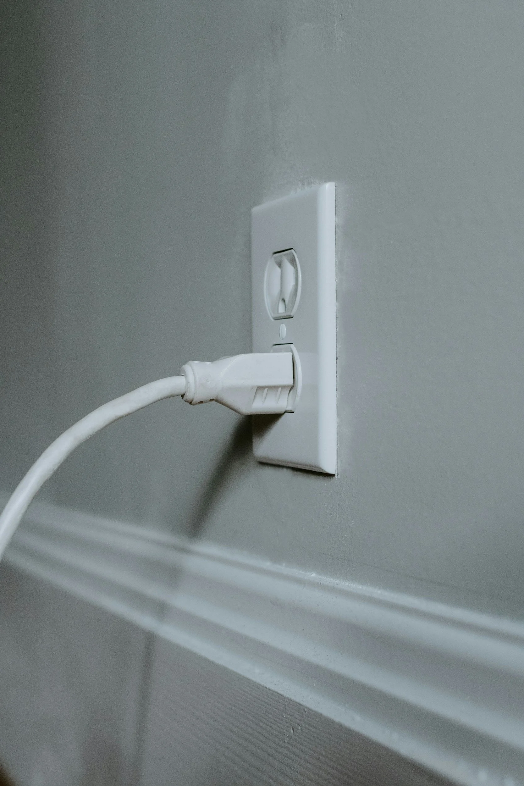 Close-up of a white electrical outlet with a connected white power cord, mounted on a light gray wall.
