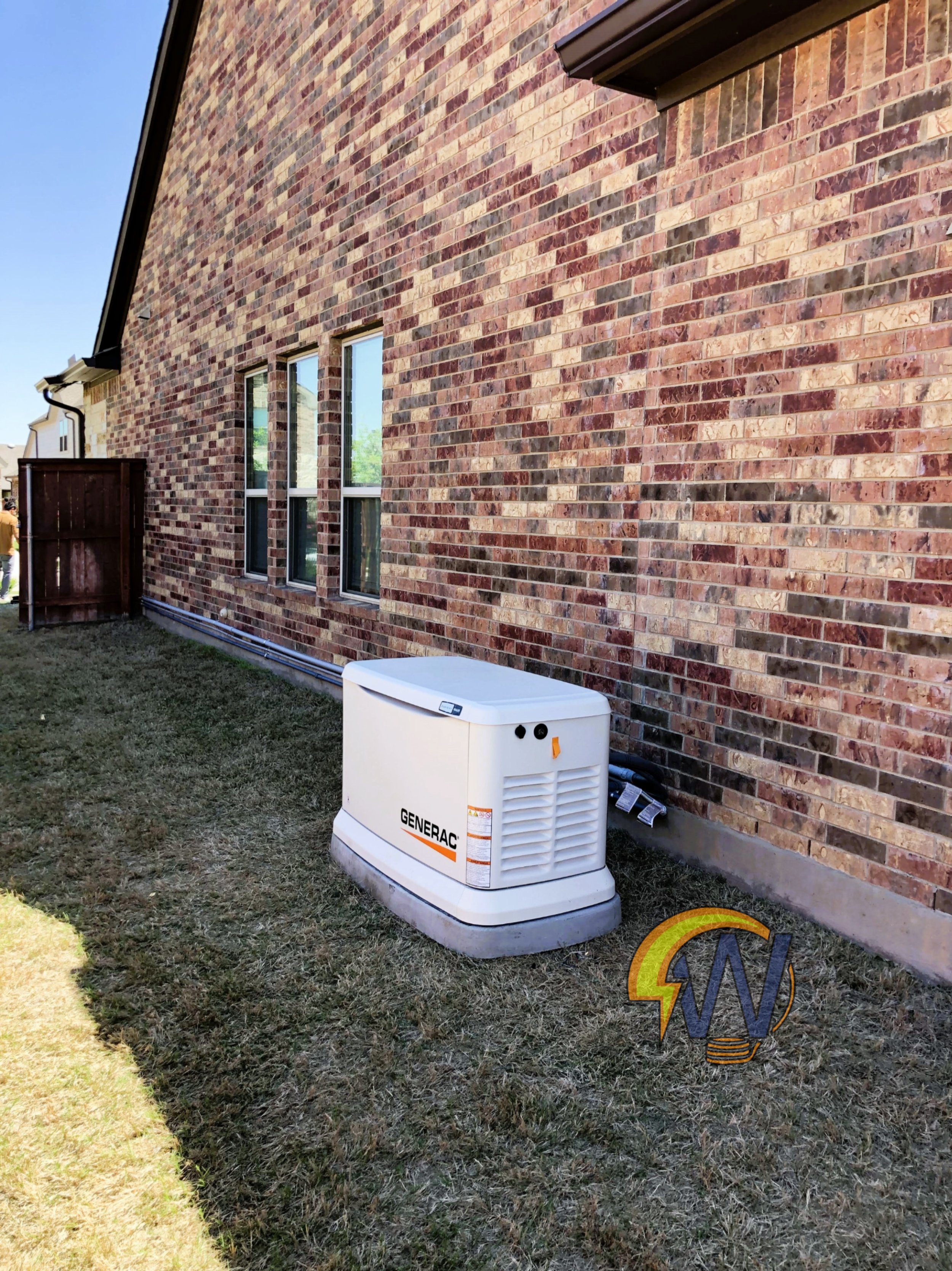 A brick house exterior with three windows, a wooden fence, a white generator unit on the ground, and a garden hose on the grass.