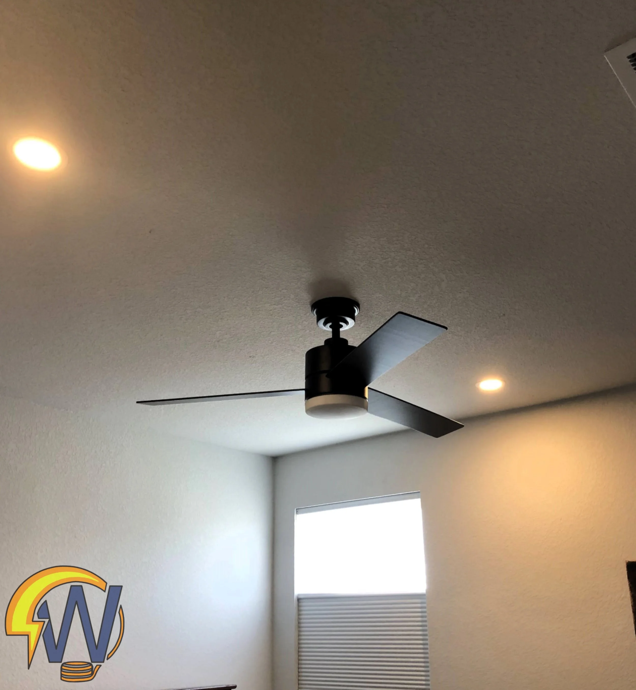 Interior view of a room with a ceiling fan and two lights, a window with blinds, part of a wall with a logo, and a white wall.