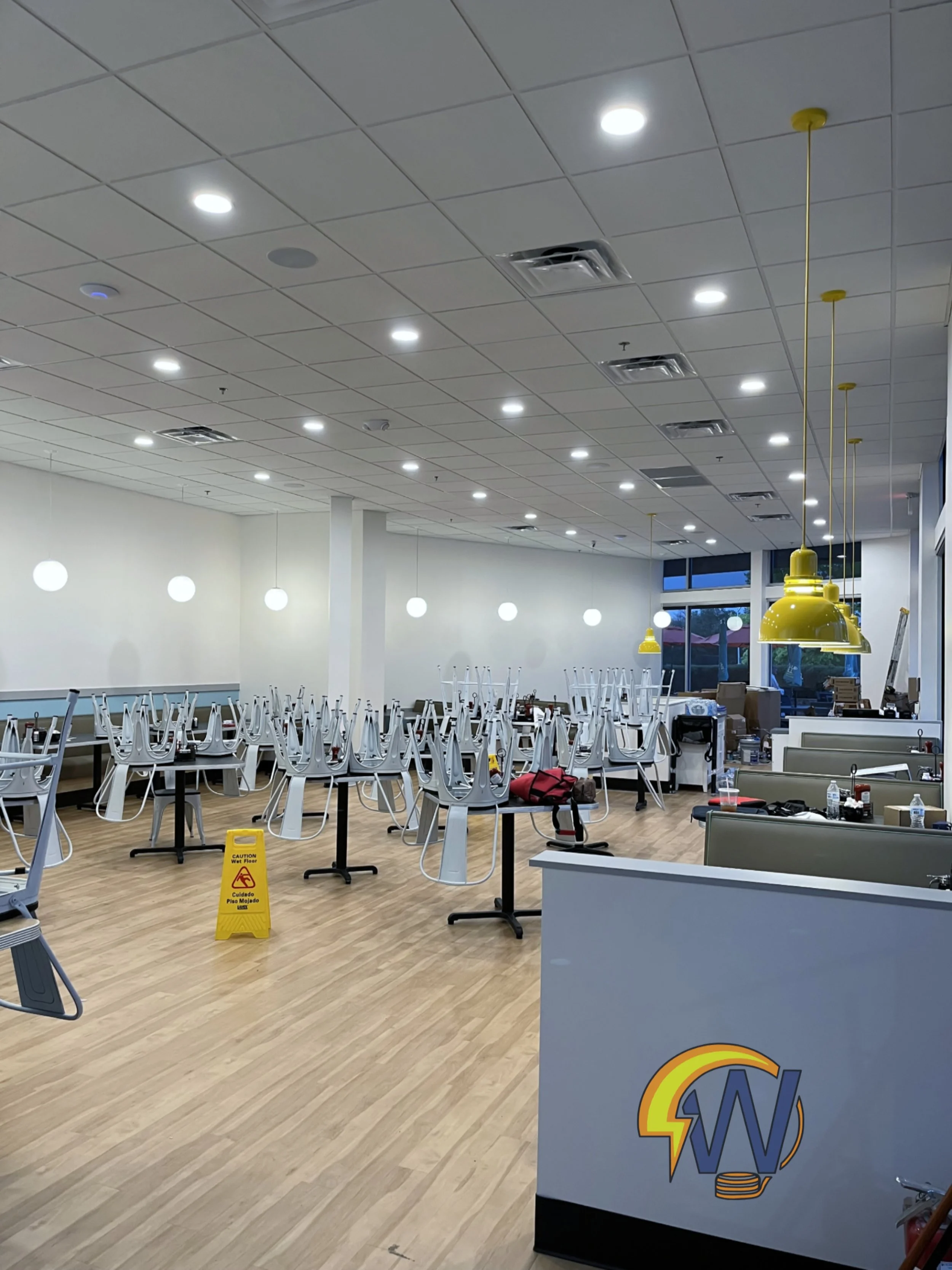 An empty restaurant with chairs stacked on tables and a caution wet floor sign, indicating it is under renovation or cleaning. The ceiling has multiple lights and ventilation units, and there are large windows on one side letting in natural light. 