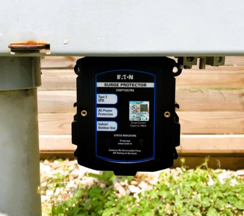 An outdoor EATON surge protector mounted under a metal surface, with green plants and wooden deck in the background.