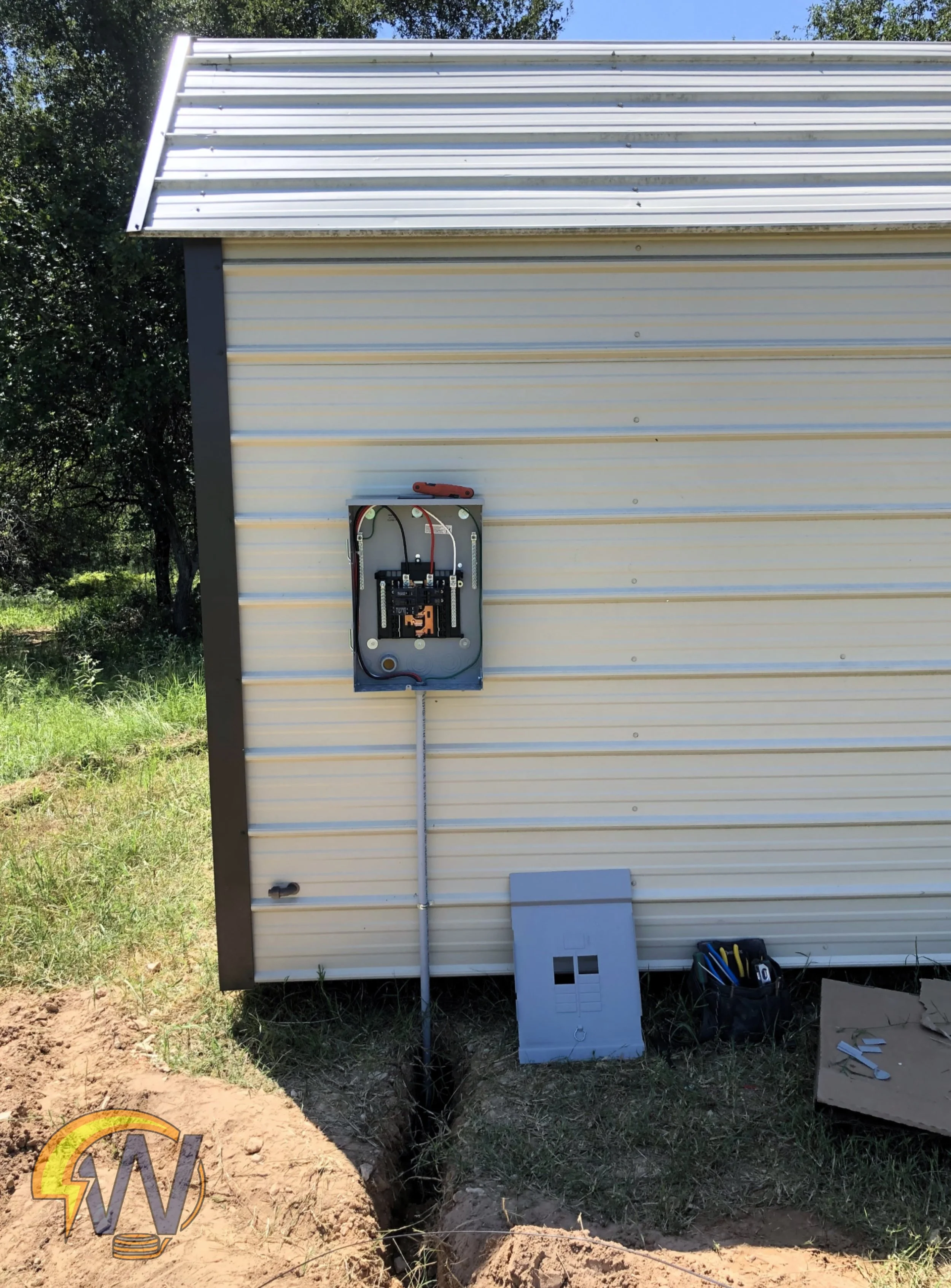The side of a building with beige siding, an electrical panel with wires, and various tools sitting on the ground nearby. There is a trench dug in the earth beneath the building, with a black pipe running through it. Toolbox and papers are on the gro