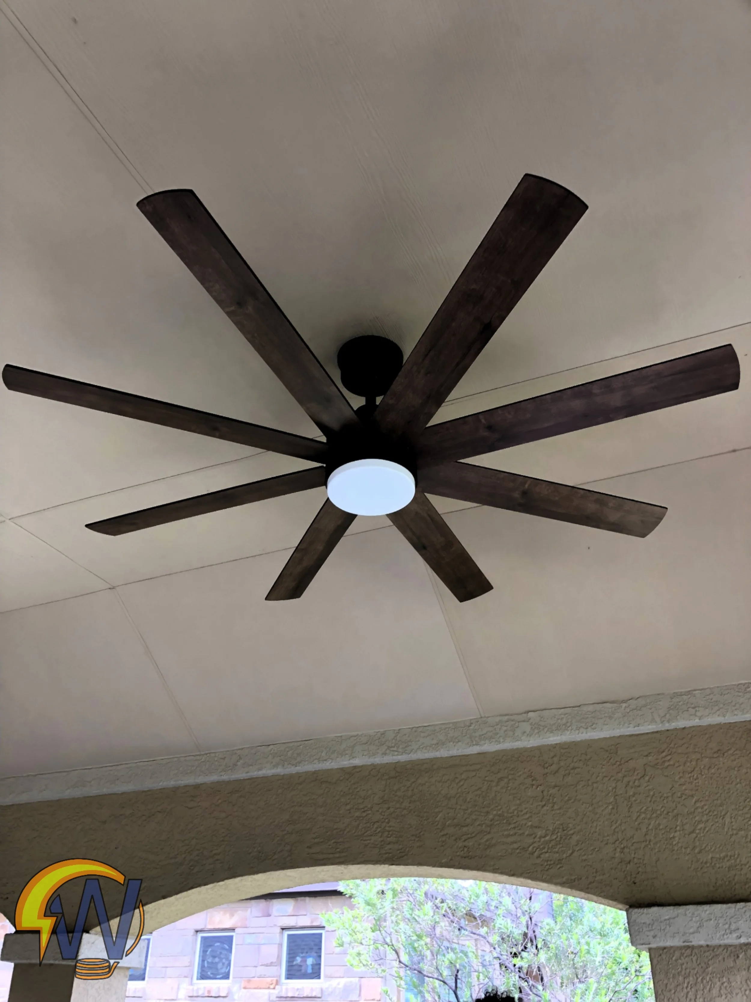 Ceiling fan with dark wooden blades and a central white light fixture mounted on a beige ceiling in an outdoor patio area, view of a house with windows and greenery outside.