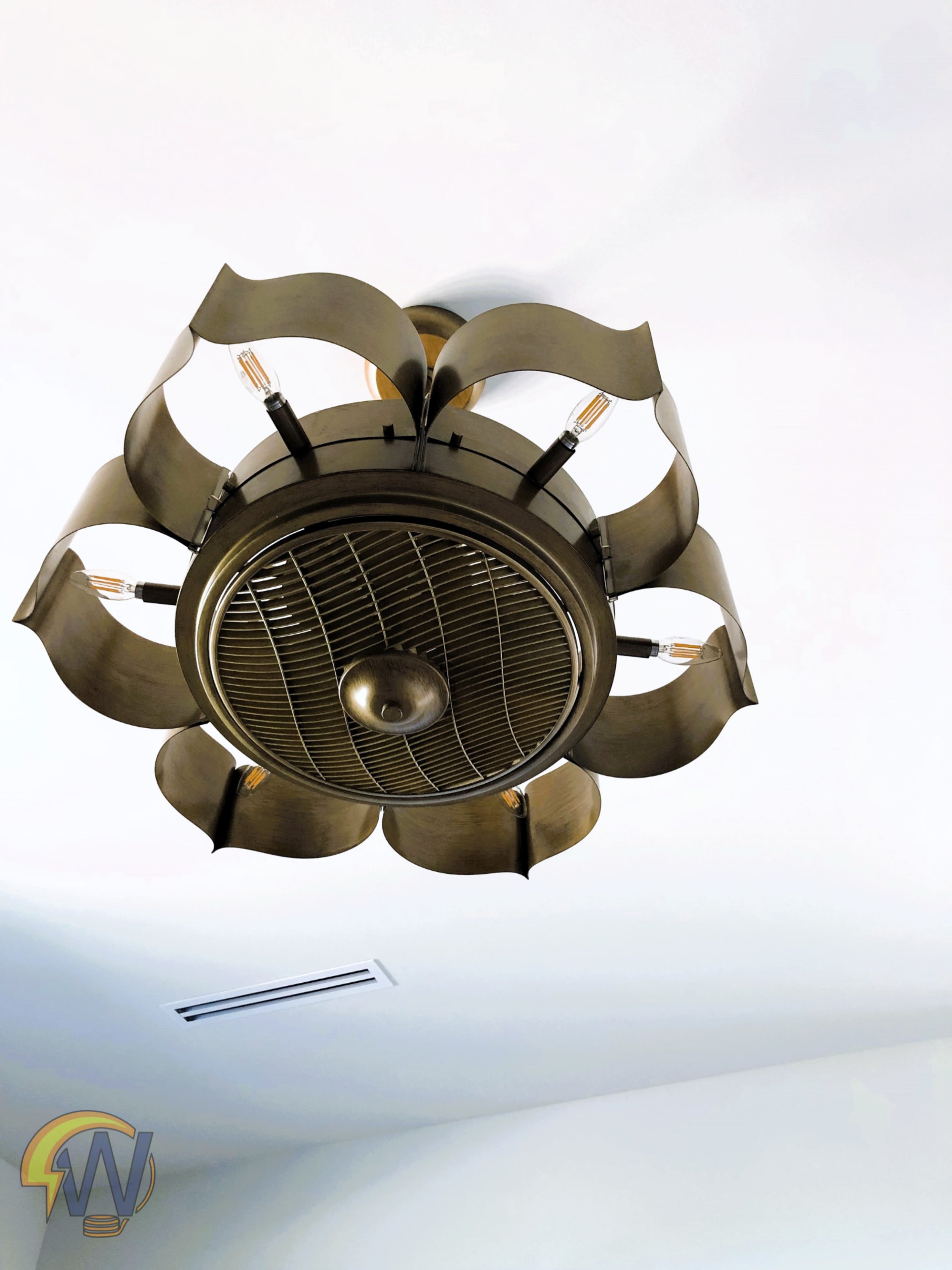 Close-up of a modern ceiling fan with a metallic finish, featuring a decorative scalloped edge around the light fixture, mounted on a white ceiling with an air vent visible nearby.