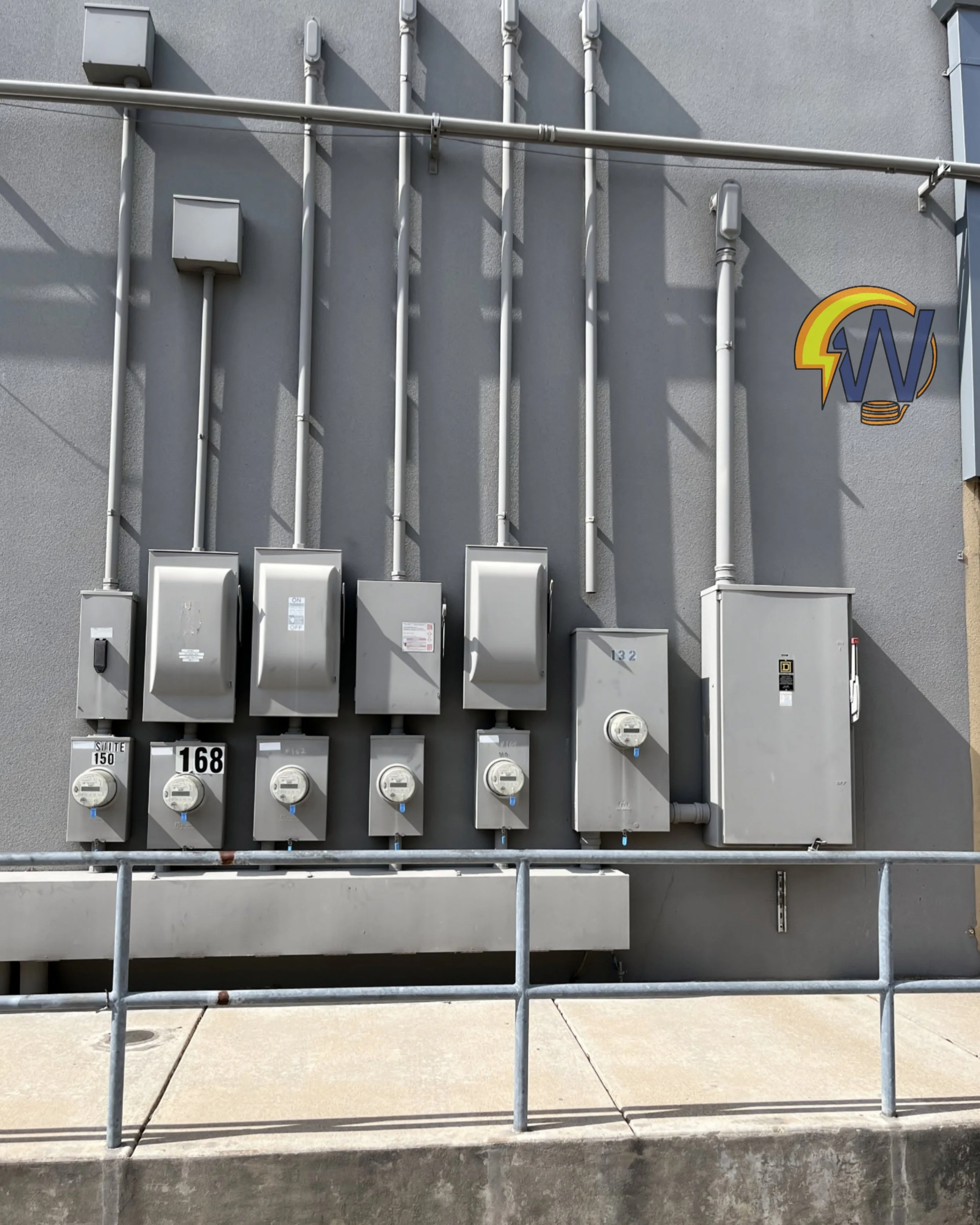 A row of electrical meters and electrical boxes mounted on a gray exterior wall, with pipes and conduit running vertically and horizontally. There is a railing in front of the setup and a concrete sidewalk below.