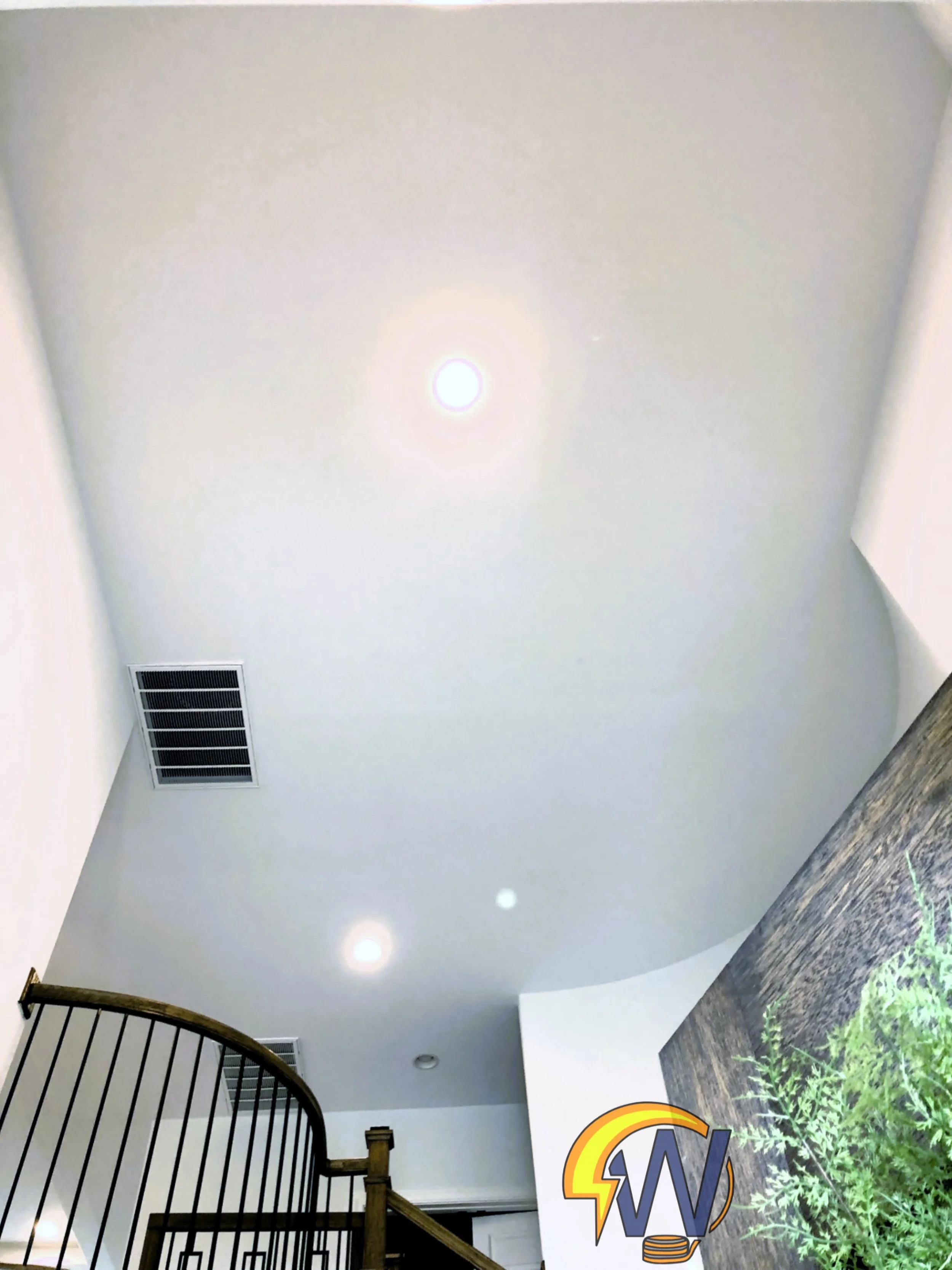 A view of a ceiling with a round light fixture, an air vent, part of a staircase with black railing, and a partial logo with colorful text and a plant on the floor.