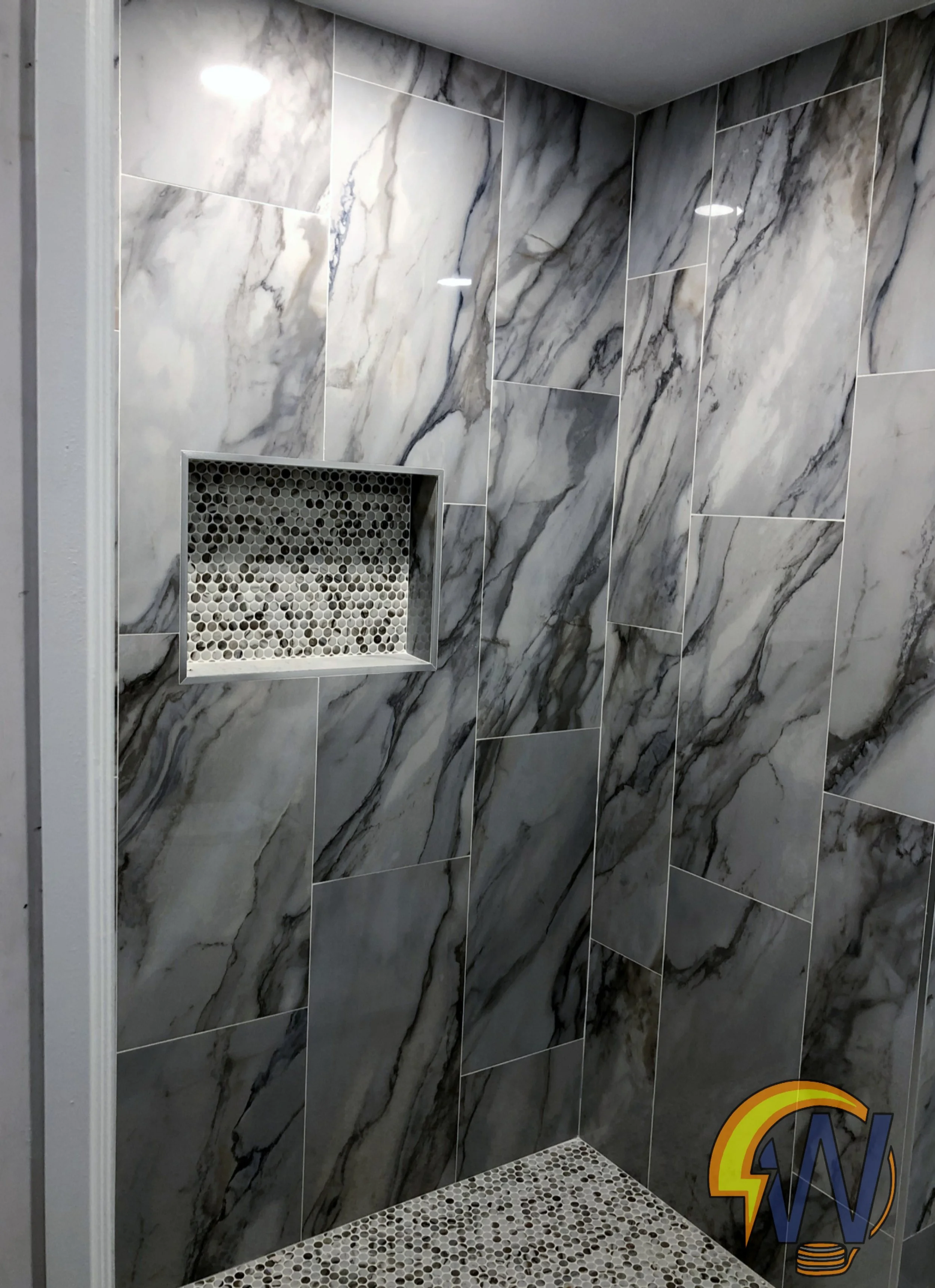 A shower with large marble tiles on the walls and a recessed shelf with a mosaic tile back. The floor has small pebble-like tiles.
