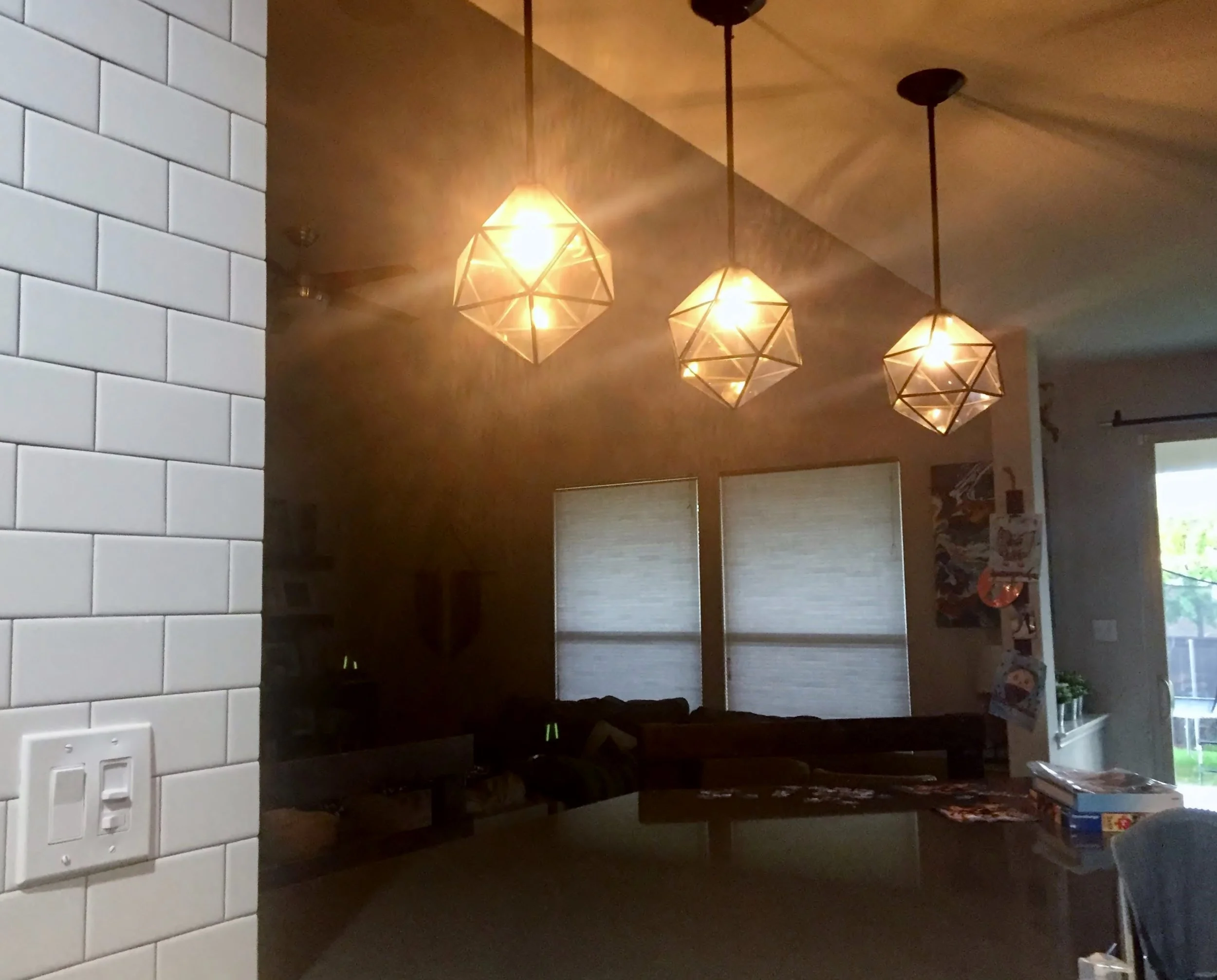 A cozy interior with three geometric pendant lights hanging from the ceiling, a white tiled wall on the left, a window with blinds on the right, and a wooden dining table with chairs and some items on it.