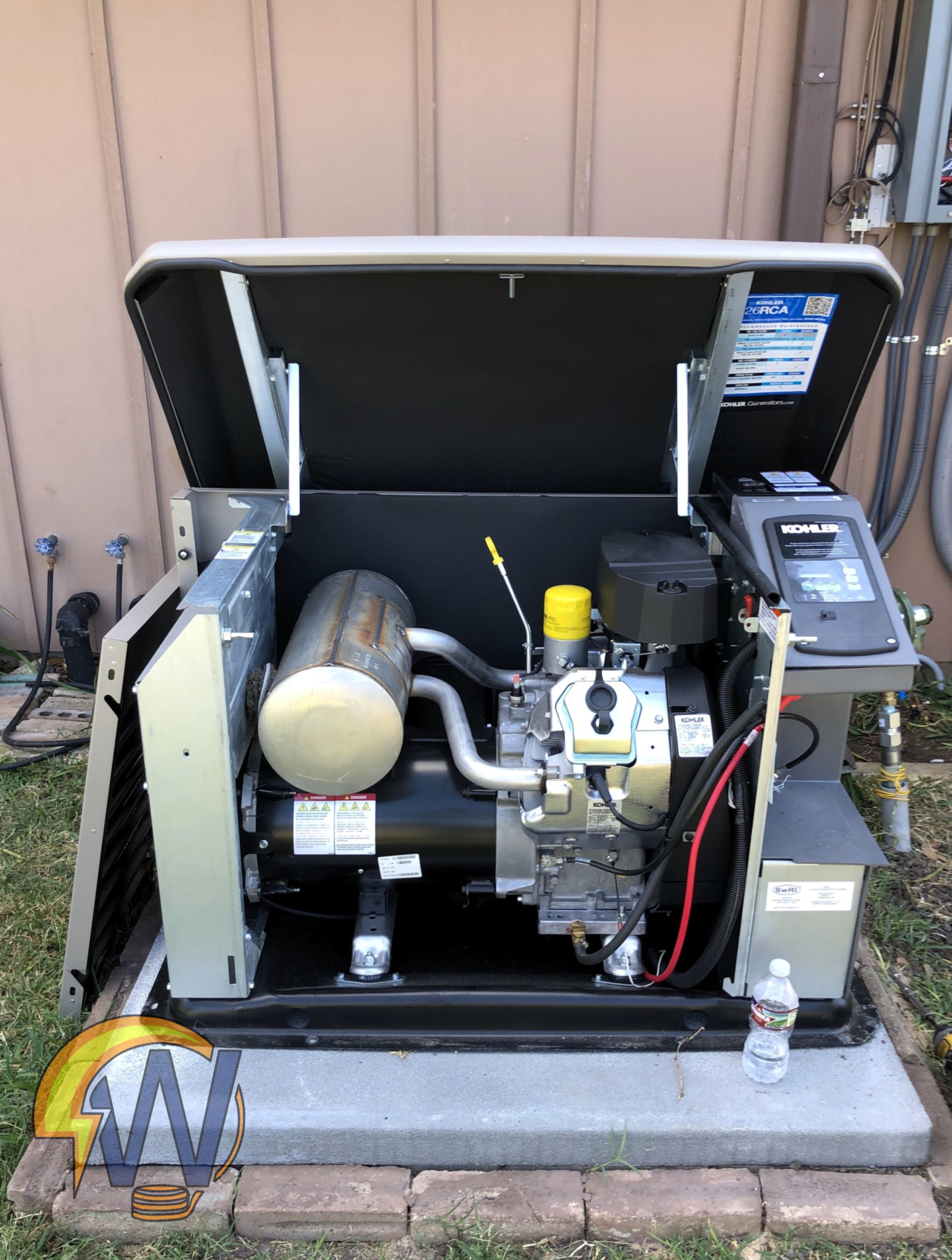 Open industrial portable generator with exposed internal parts, located outdoors on a concrete and brick platform next to a beige building wall, with a plastic water bottle on the ground nearby.