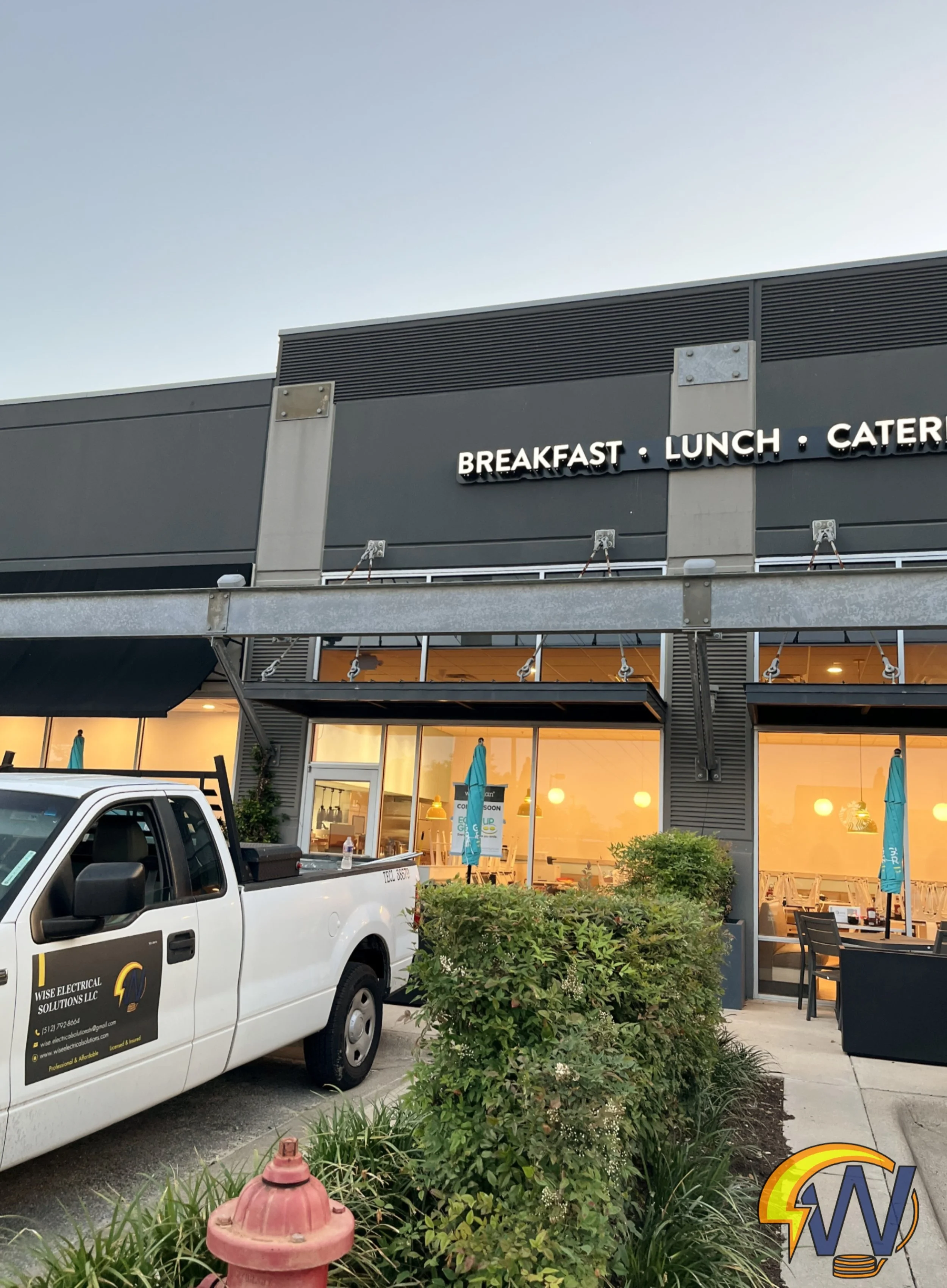 The image shows the exterior of a modern building with a sign that reads 'Breakfast, Lunch, Catering'. There is a white truck parked in front, and the building has large windows with interior lighting, outdoor seating with umbrellas, and some greener