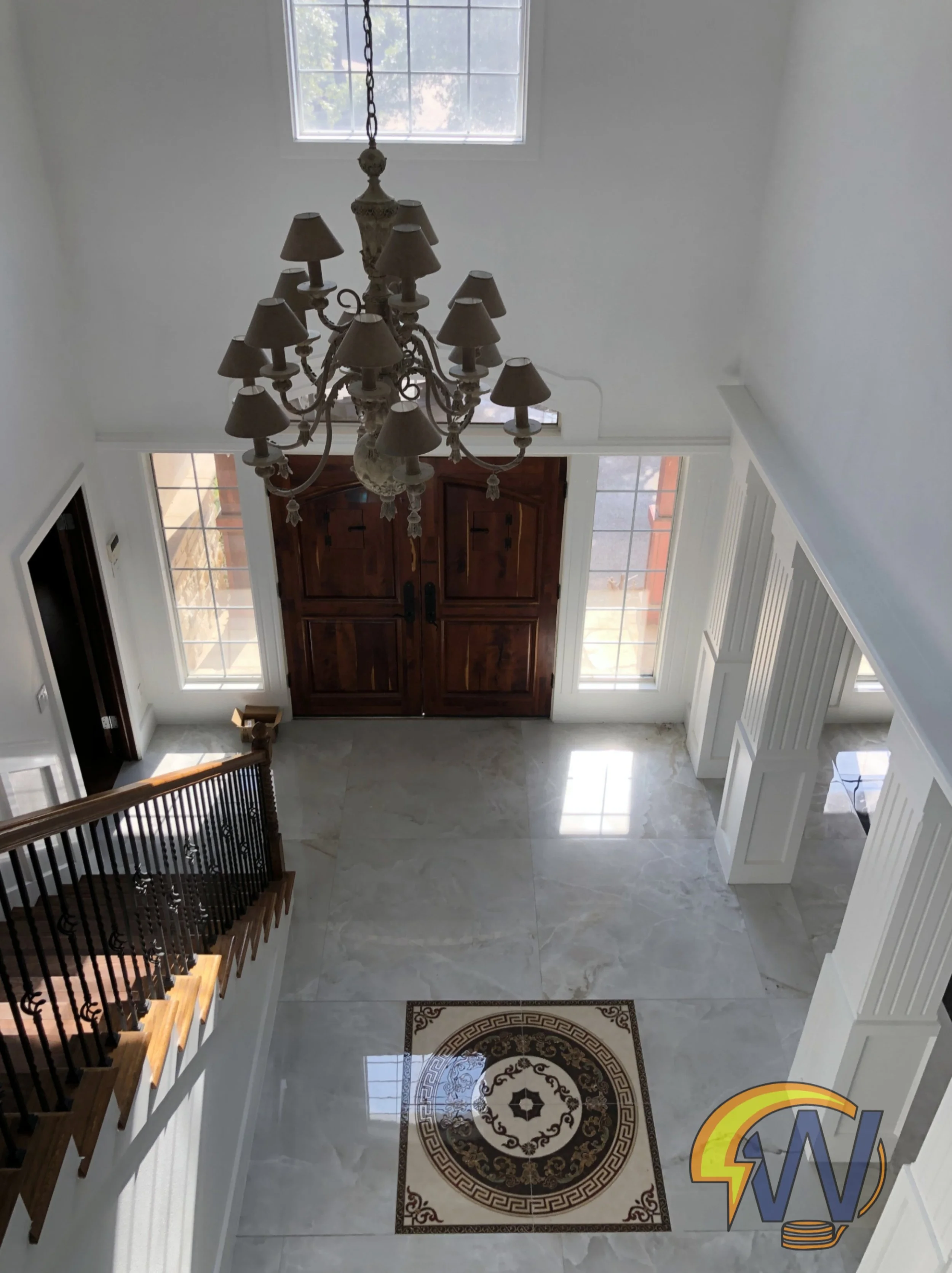 A view of a home's entryway with tiled flooring, a decorative rug, wooden front door, large windows, and a chandelier hanging from the ceiling.