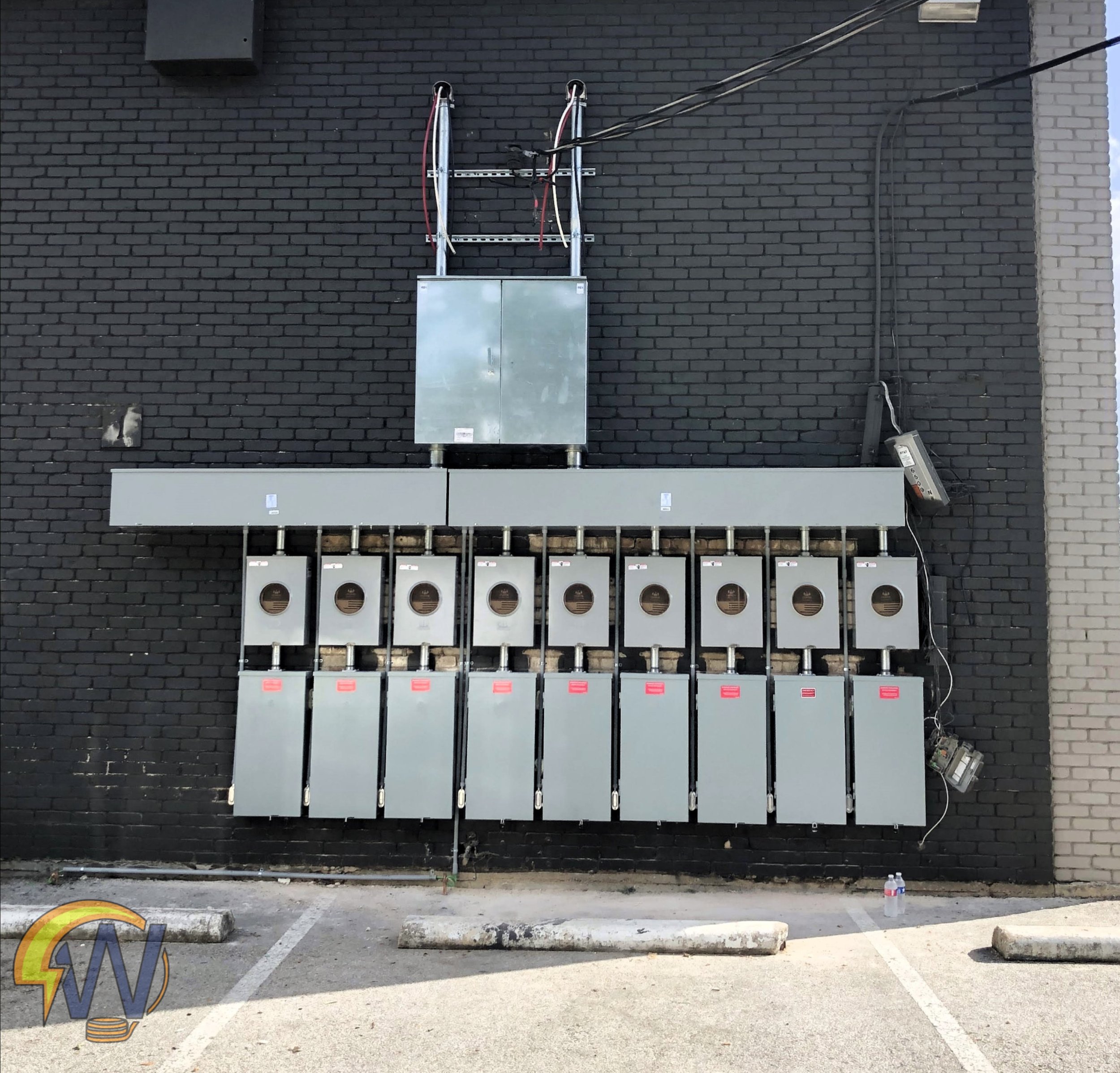 Electrical boxes and wiring mounted on a black brick wall in a parking lot with concrete wheel stops and traffic cones.