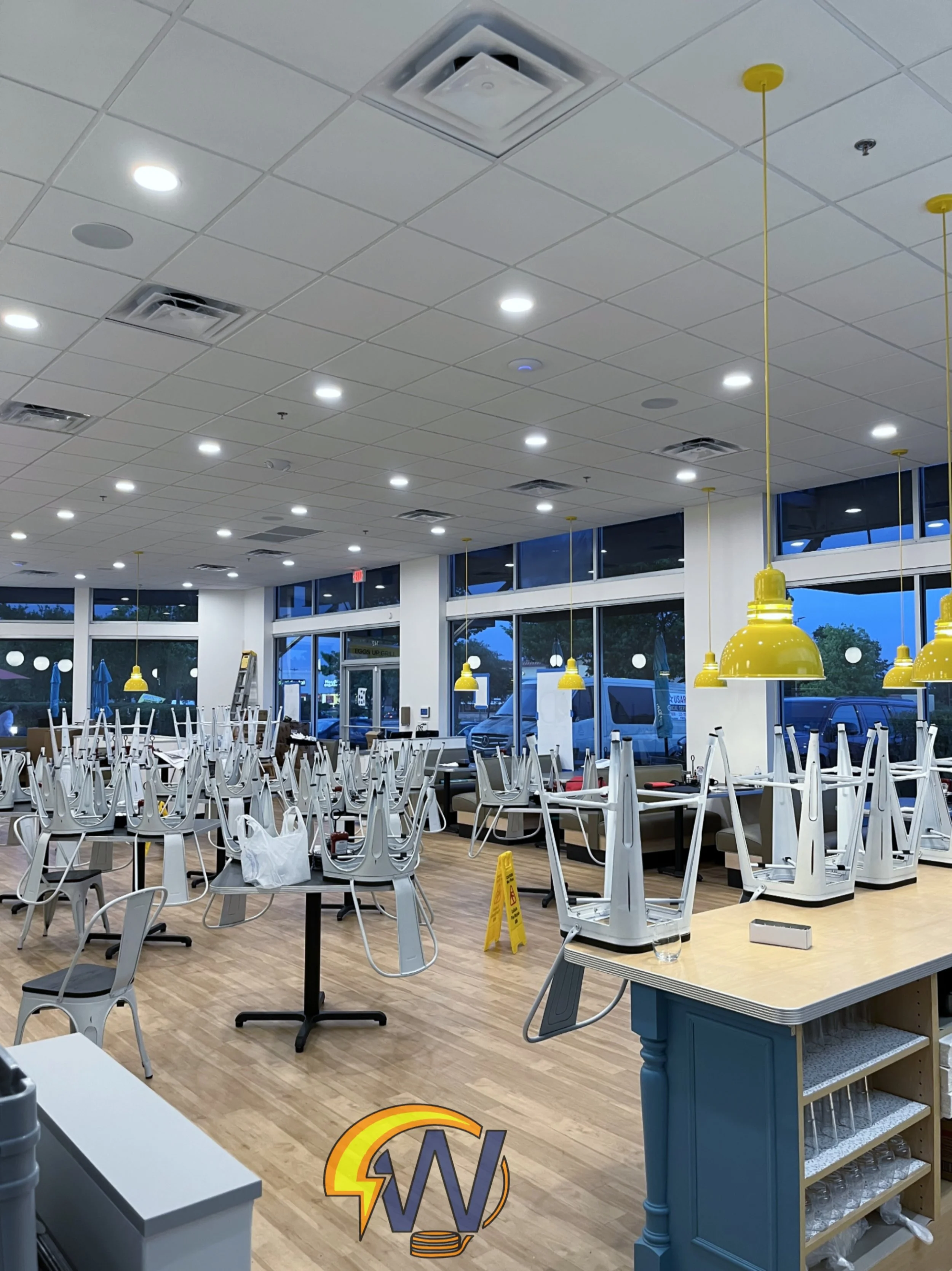 Interior of a restaurant or café with chairs stacked upside down on tables, yellow hanging lamps, large windows, and mopping sign on the floor.