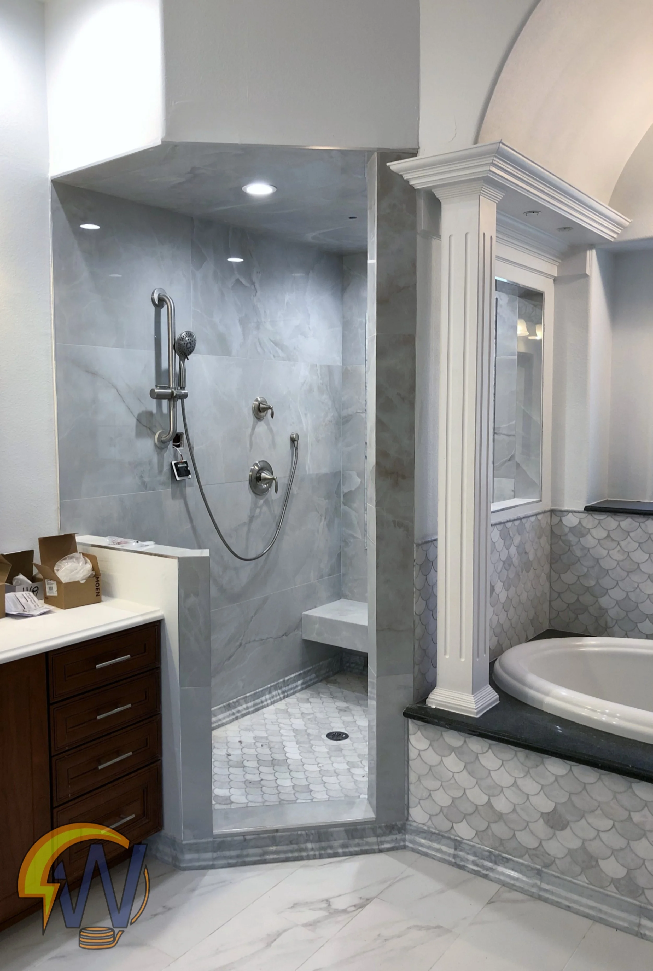 A bathroom display with a shower area featuring marble tiles, a rainfall showerhead, and built-in bench, next to a decorative column with a mirror above the bathtub with light-colored fish scale tiles on the wall.